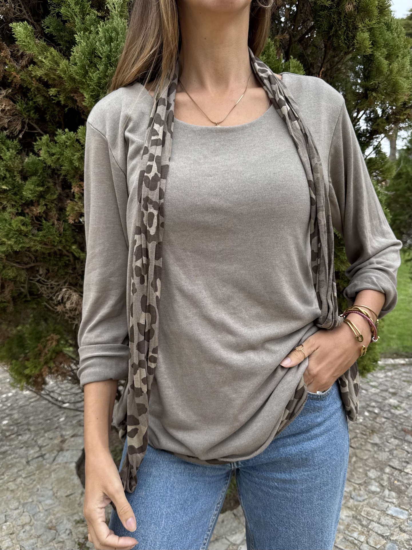 Mulher com camisola cinza, cachecol animal print, jeans azul e pulseiras, em exterior com vegetação e chão de calçada