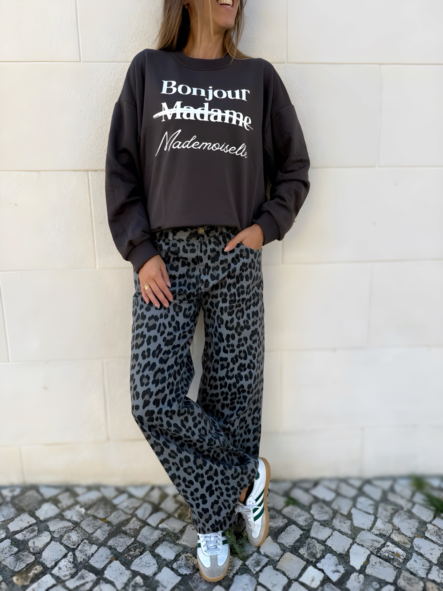 Mulher com camisola preta com texto, calças animal print e sapatos brancos numa calçada portuguesa