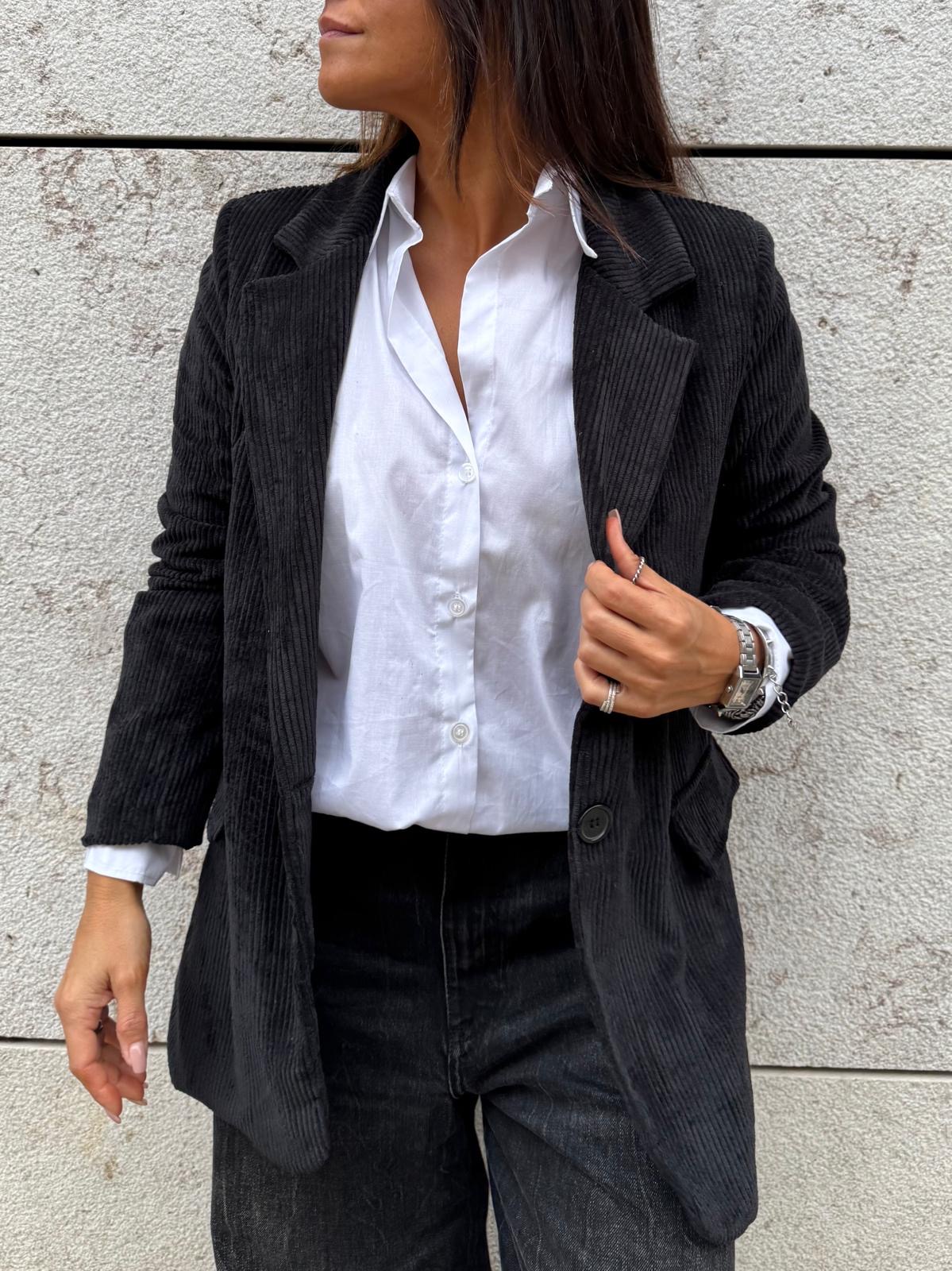 Blazer preto de veludo cotelê com camisa branca e calças pretas em fundo de parede de pedra clara