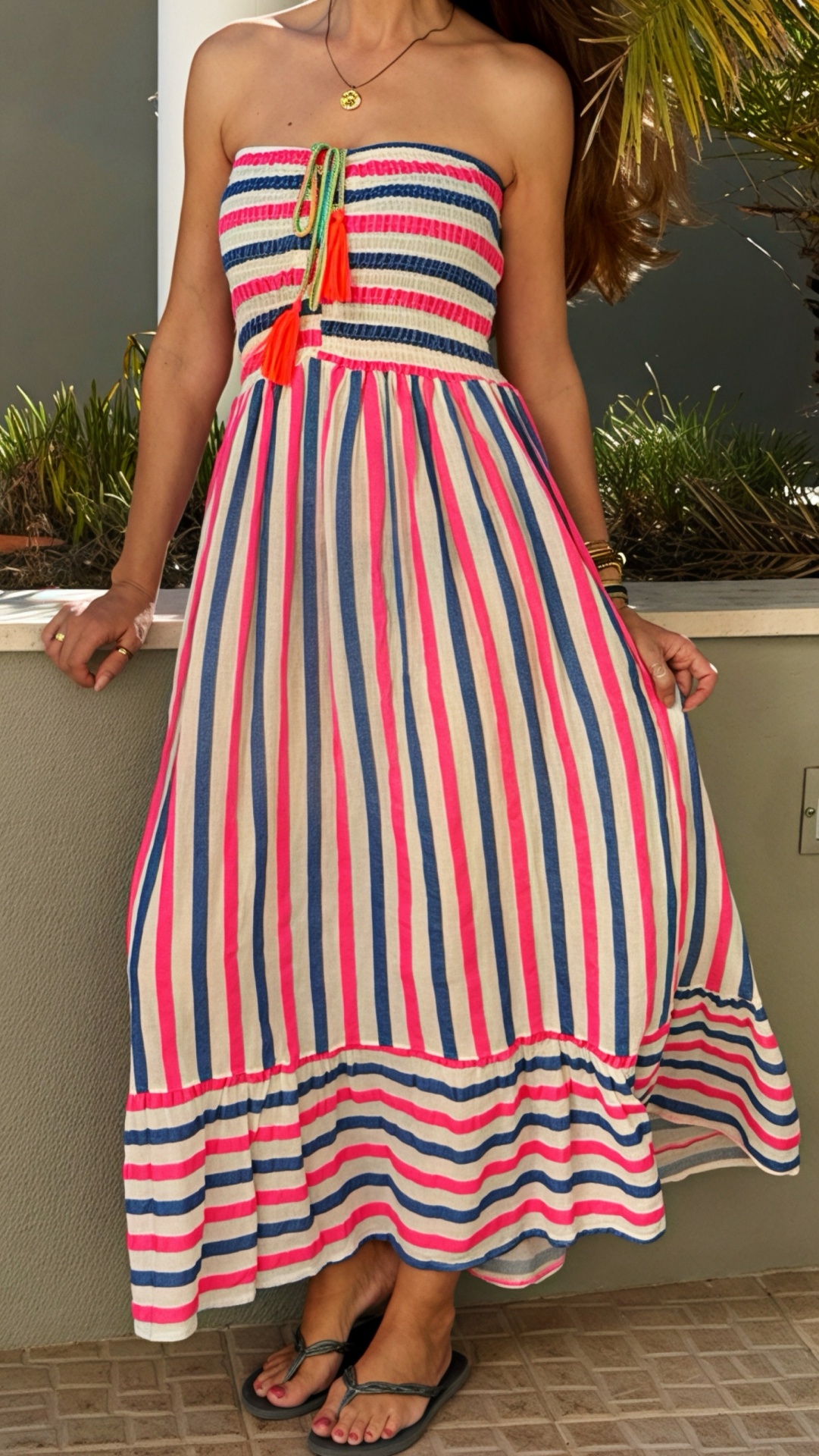 Vestido sem alças com riscas azuis, rosa e branca e detalhe de laços verdes com tasséis laranja.