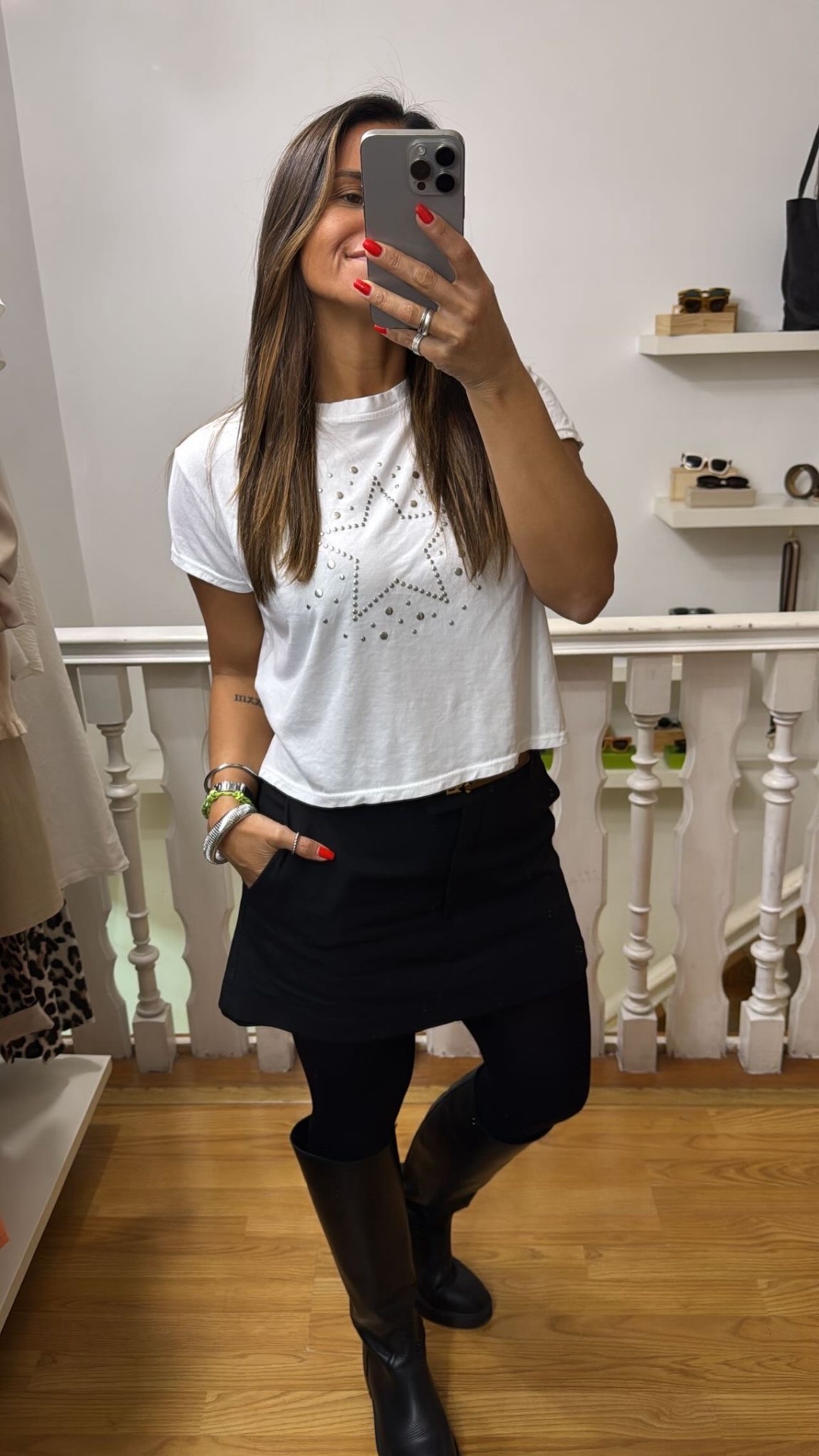 Mulher a vestir camiseta branca com estrela prateada, saia preta e botas pretas, a tirar selfie num interior com chão de madeira.