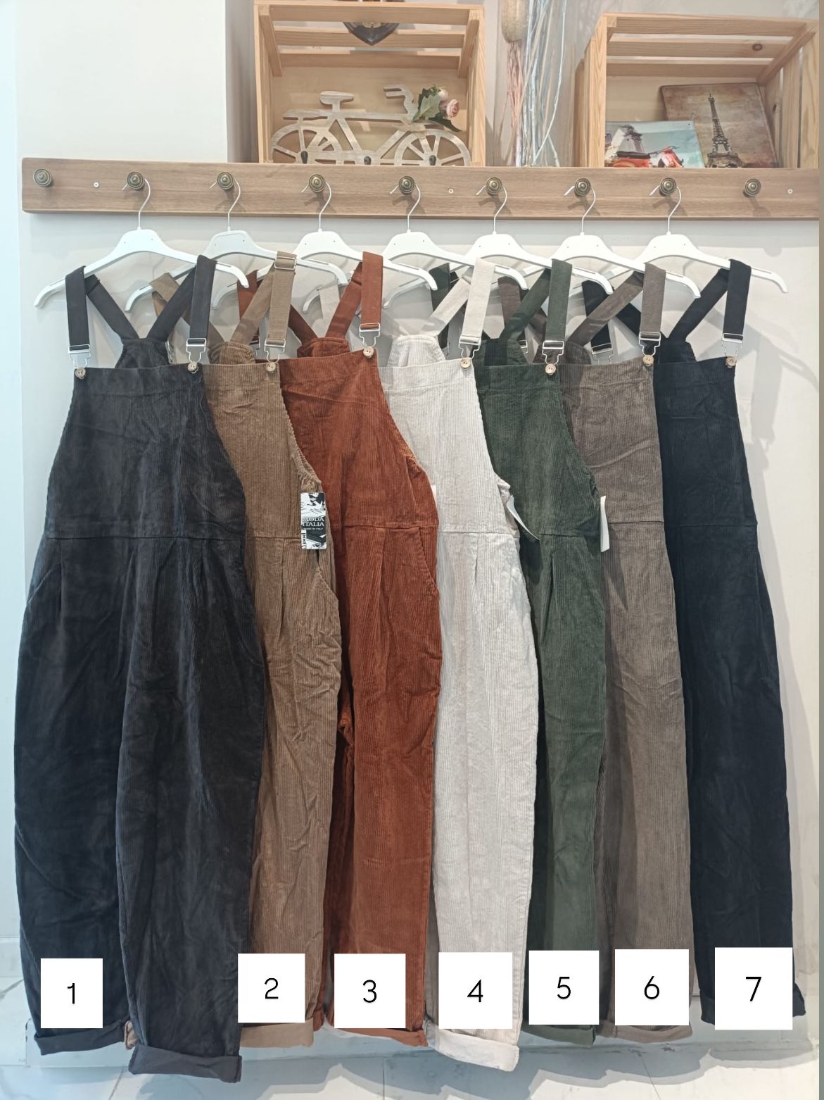 Sete jardineiras de veludo em diferentes cores penduradas numa parede branca com gancho de madeira.