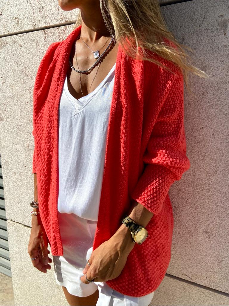 Mulher a usar cardigan vermelho e vestido branco junto a parede de pedra clara.