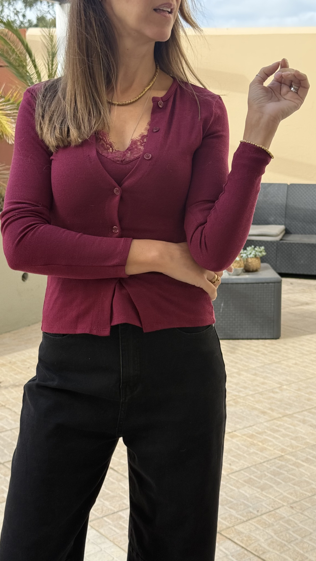 Mulher com camisola bordeaux e calças pretas no exterior, com mobília de jardim e planta ao fundo
