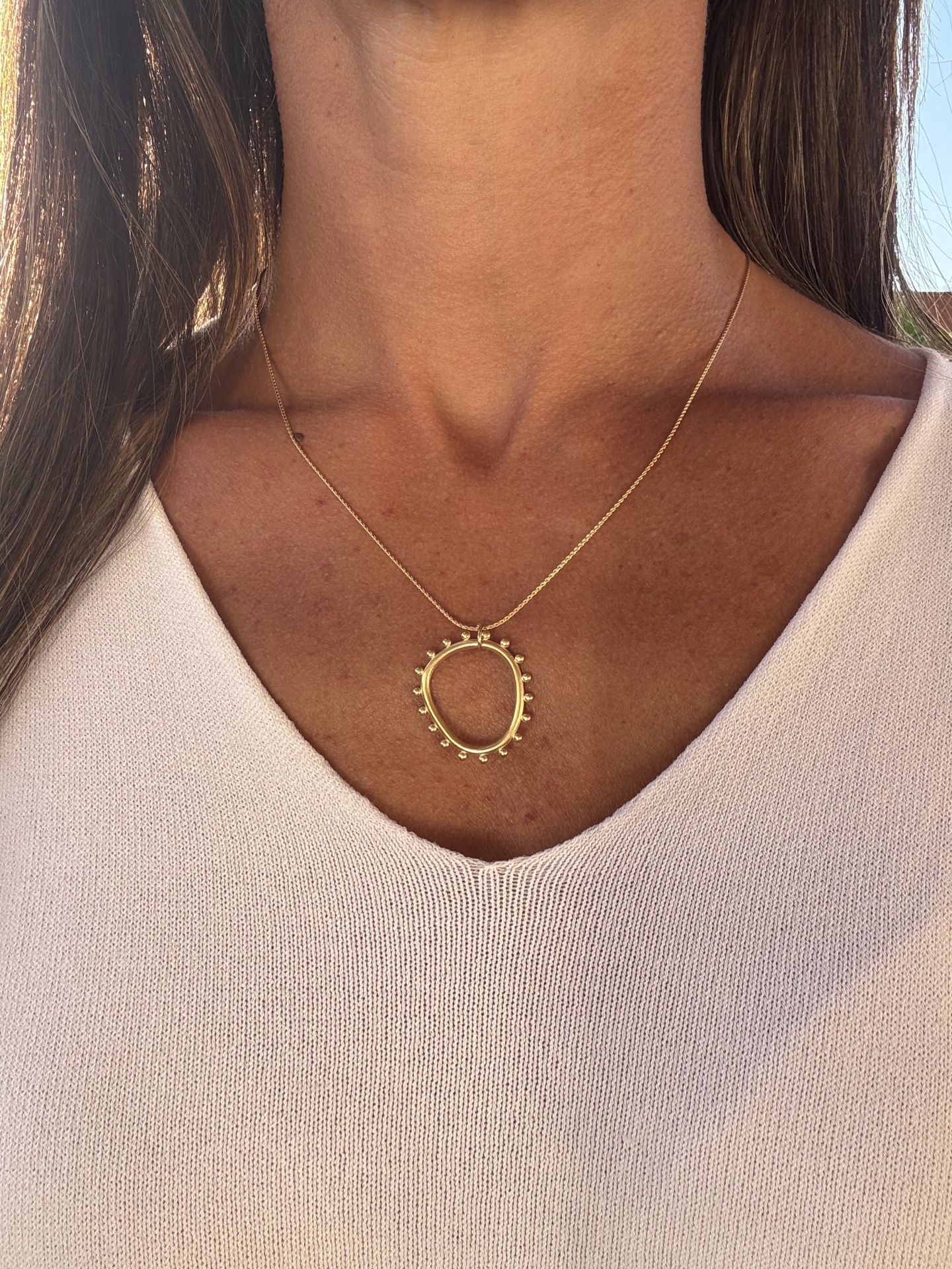 Colar dourado com pendente circular usado ao pescoço sobre camisola rosa clara