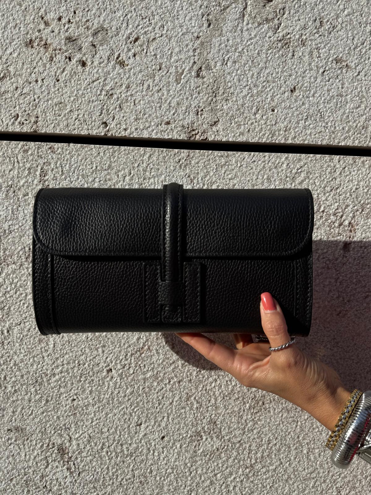 Carteira preta tipo clutch em pele com fecho, sobre fundo de parede clara, segurada por mão feminina