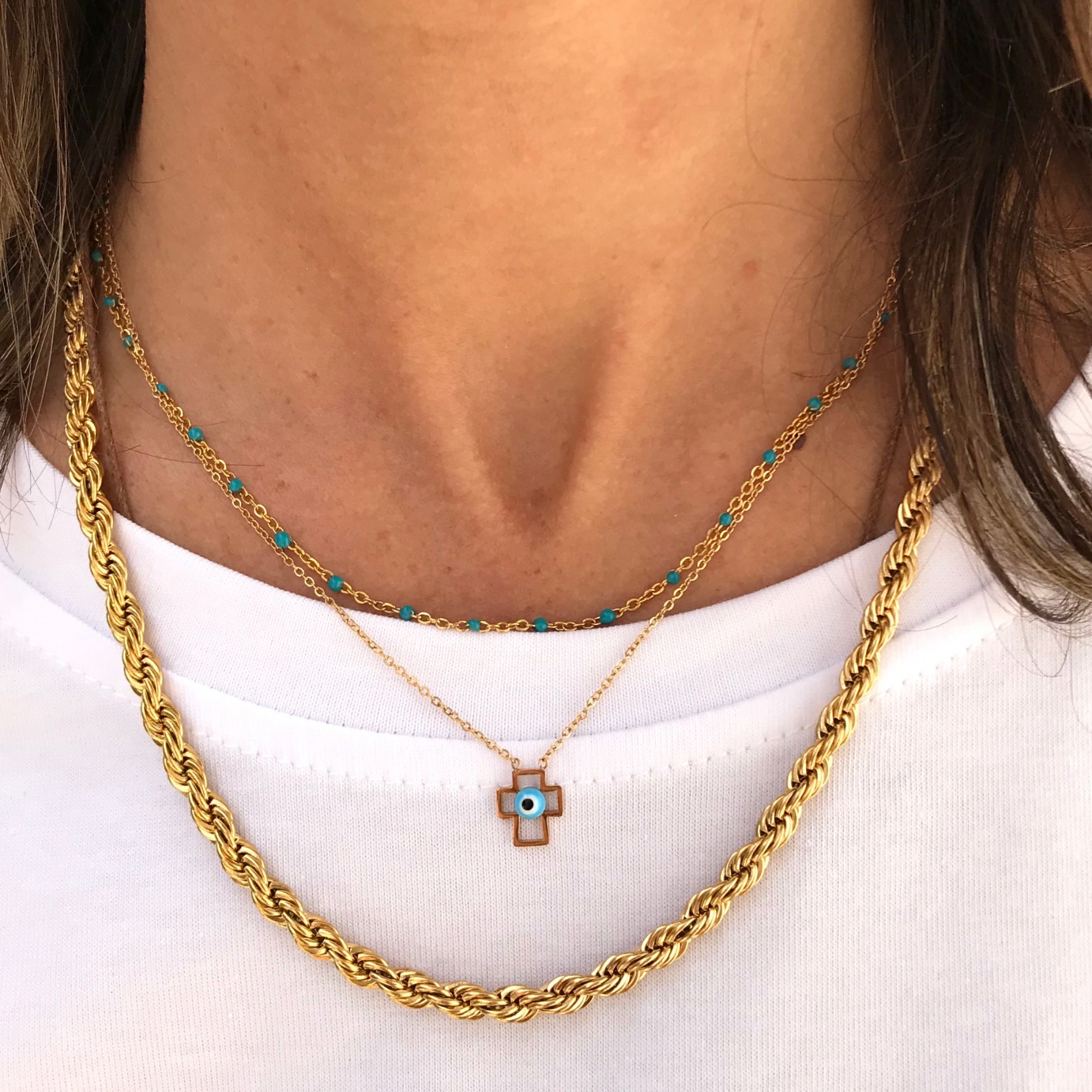 Colares dourados com corrente de pedras turquesa e pendente de cruz com olho azul sobre t-shirt branca