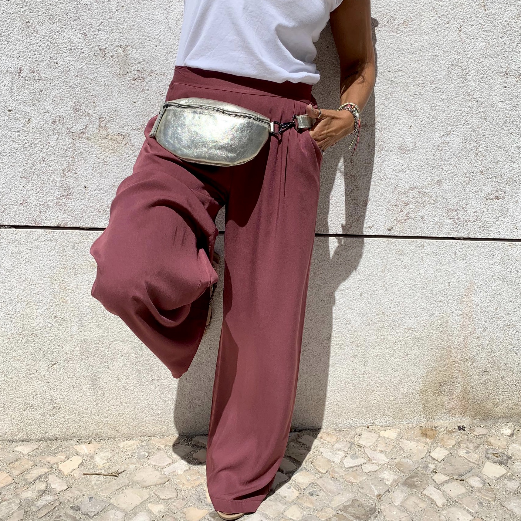 Pessoa com calças vinho largas, camiseta branca e pochete metálica prateada junto a parede branca