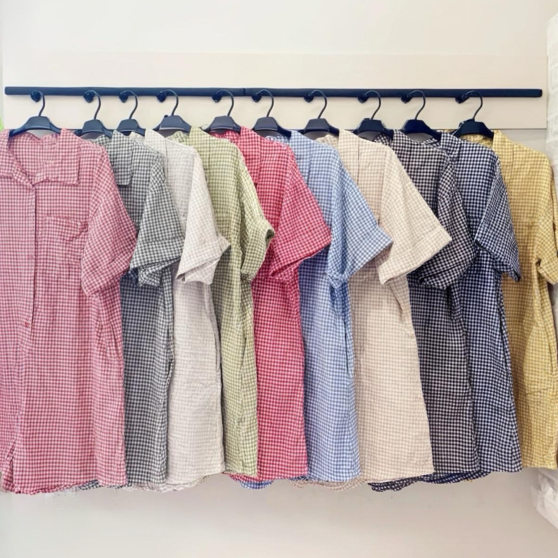 Camisas de manga curta xadrez em várias cores penduradas numa barra preta contra parede branca