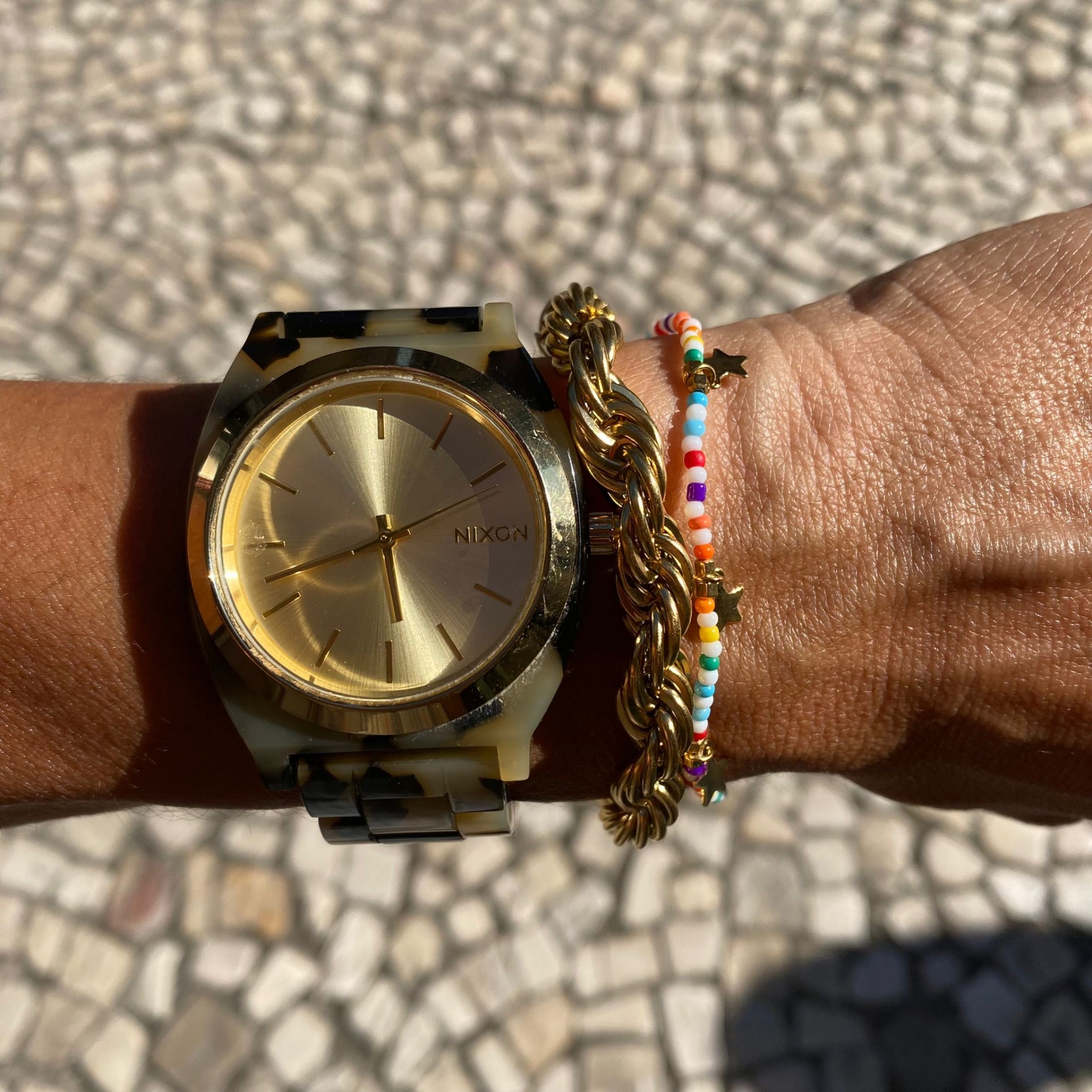 Relógio dourado Nixon com bracelete padrão tartaruga e duas pulseiras coloridas no pulso