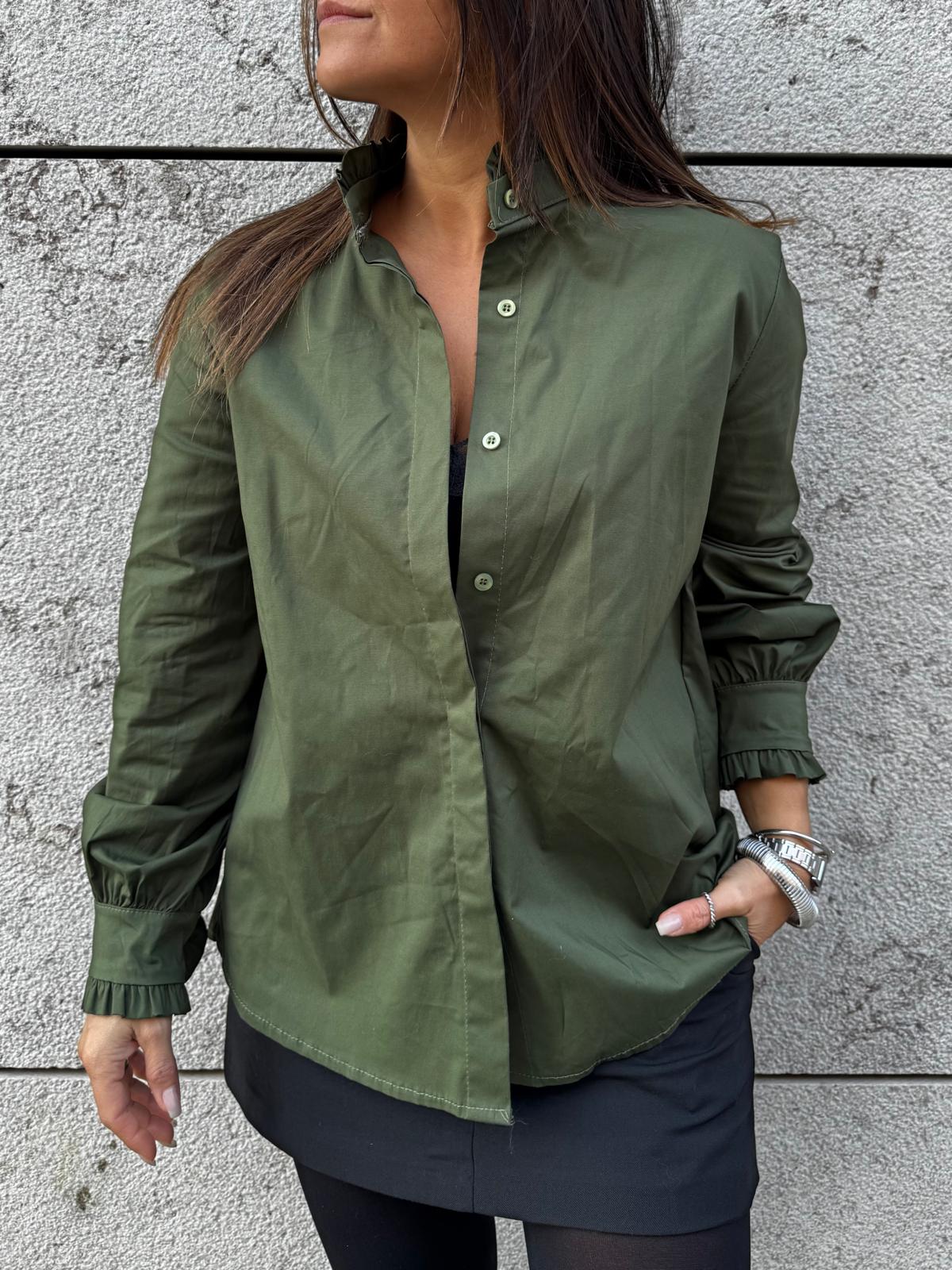 Camisa verde-oliva com botões e mangas compridas, usada por mulher com saia preta e pulseiras, parede de pedra cinza ao fundo