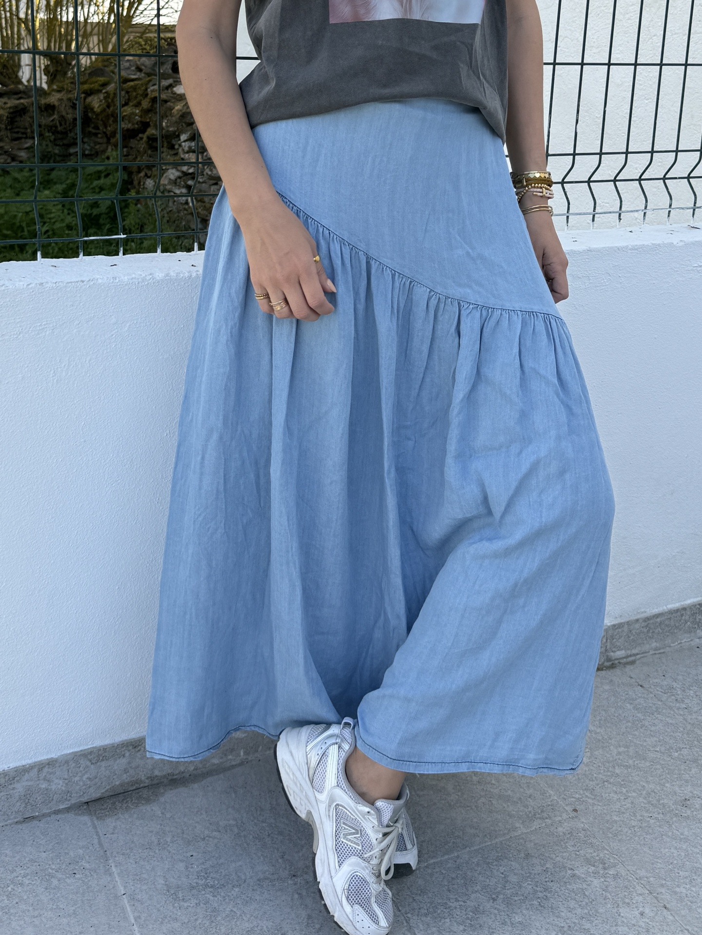Pessoa vestindo saia comprida azul claro e ténis brancos em ambiente exterior com muro branco