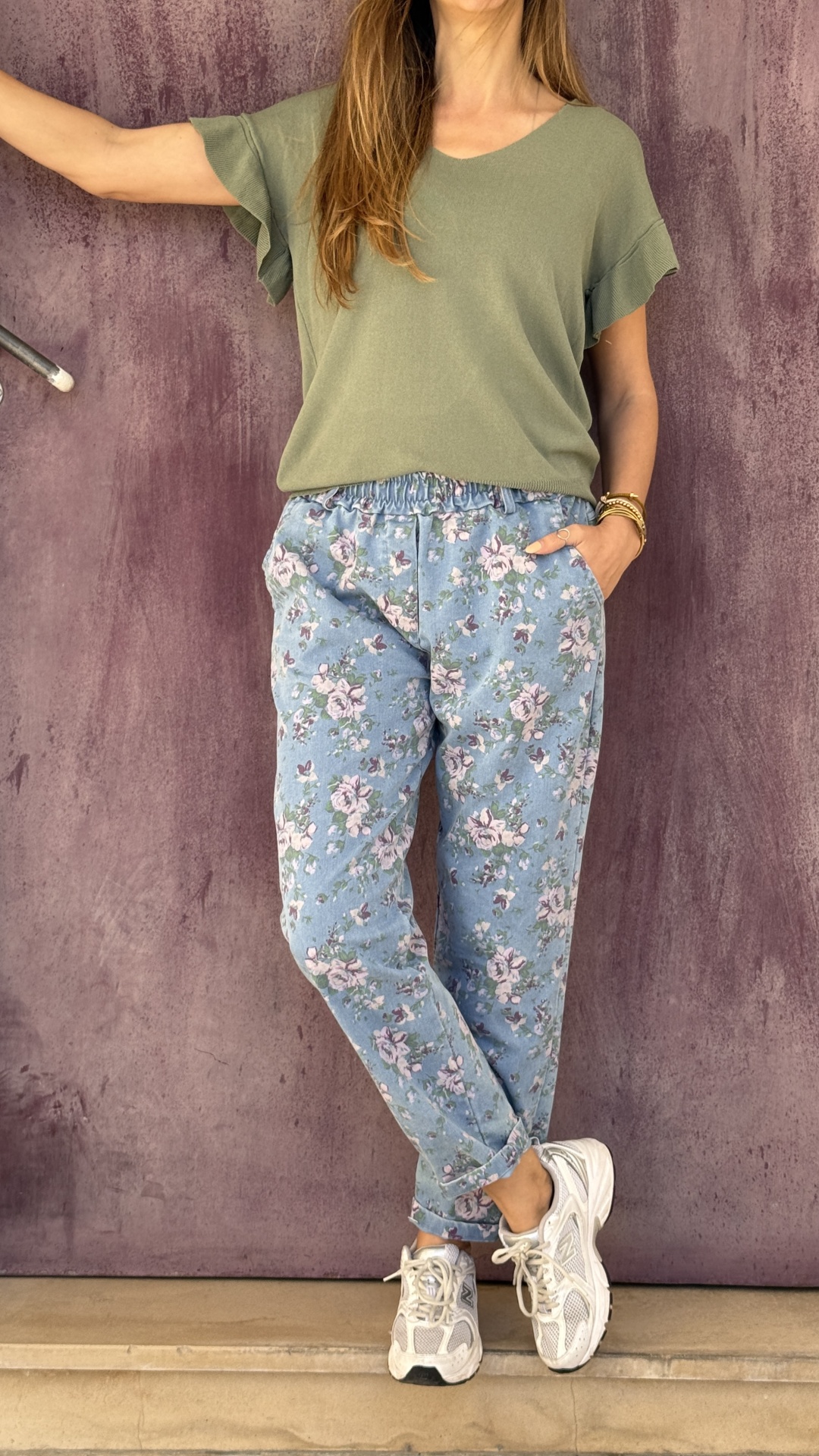 Mulher vestindo t-shirt verde, calças florais azuis claras e tênis brancos em parede roxa