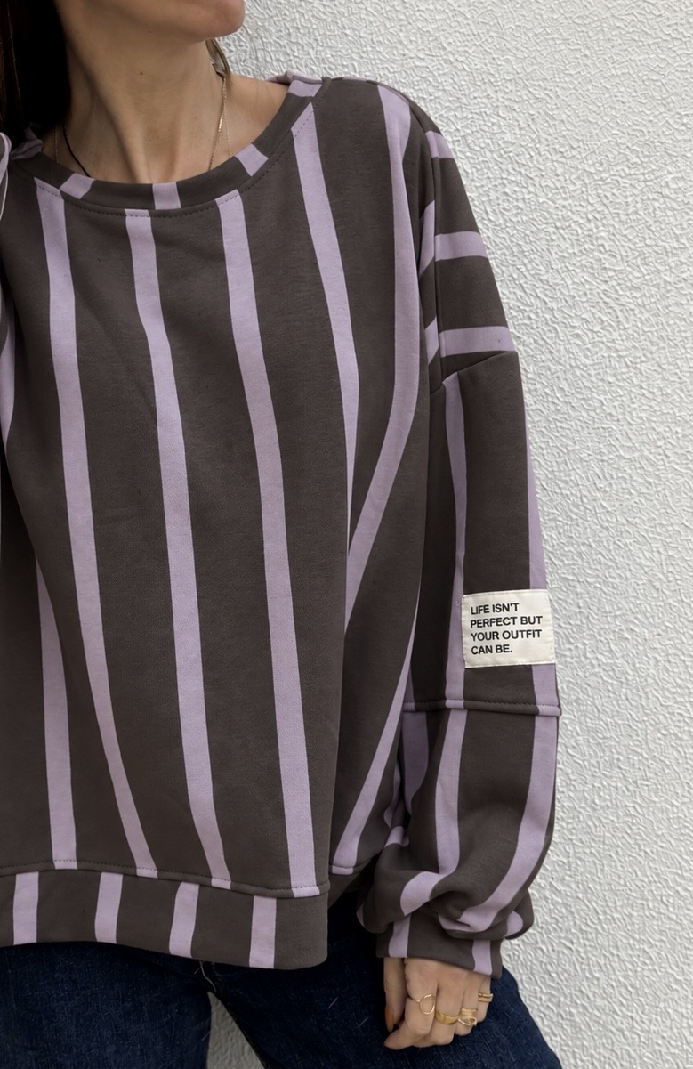 Camisola riscada lilás e castanha com etiqueta de texto na manga, usada por pessoa próxima de parede branca