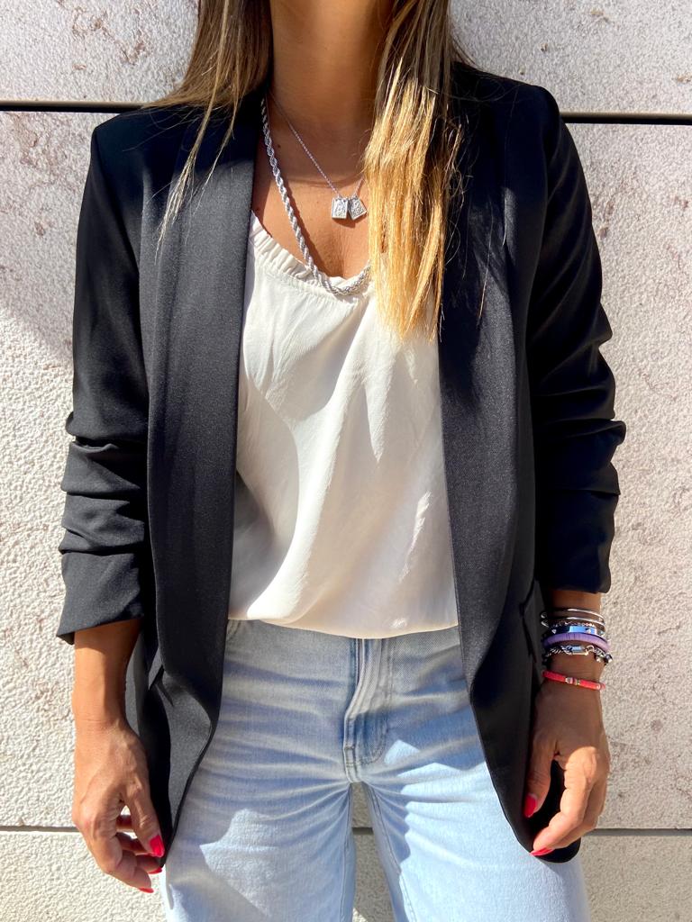 Mulher com blusa branca, blazer preto e calças de ganga azuis claras junto a parede clara