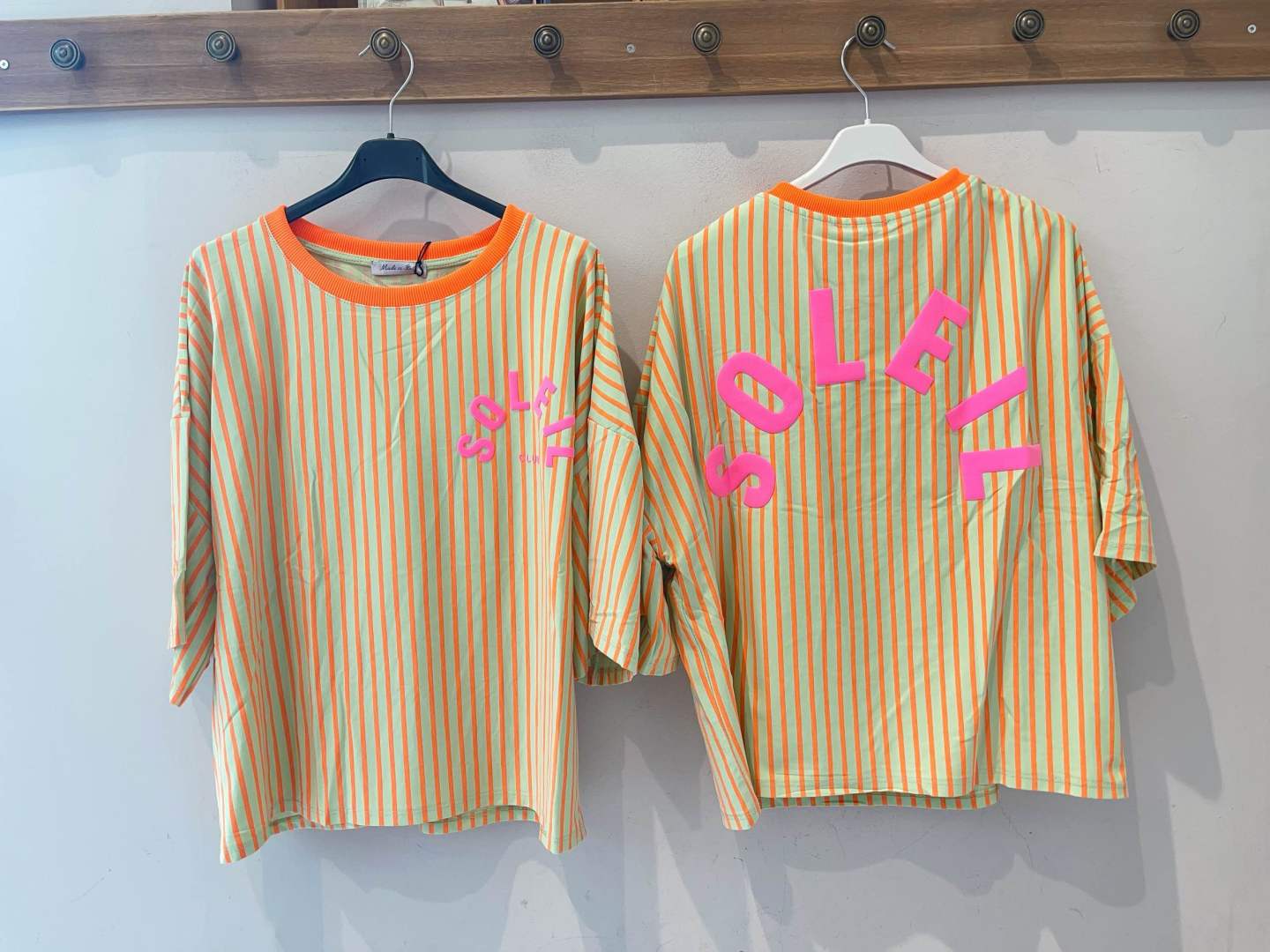 Duas t-shirts laranja com riscas verticais e a palavra SOLEIL em rosa, penduradas num cabideiro de madeira.