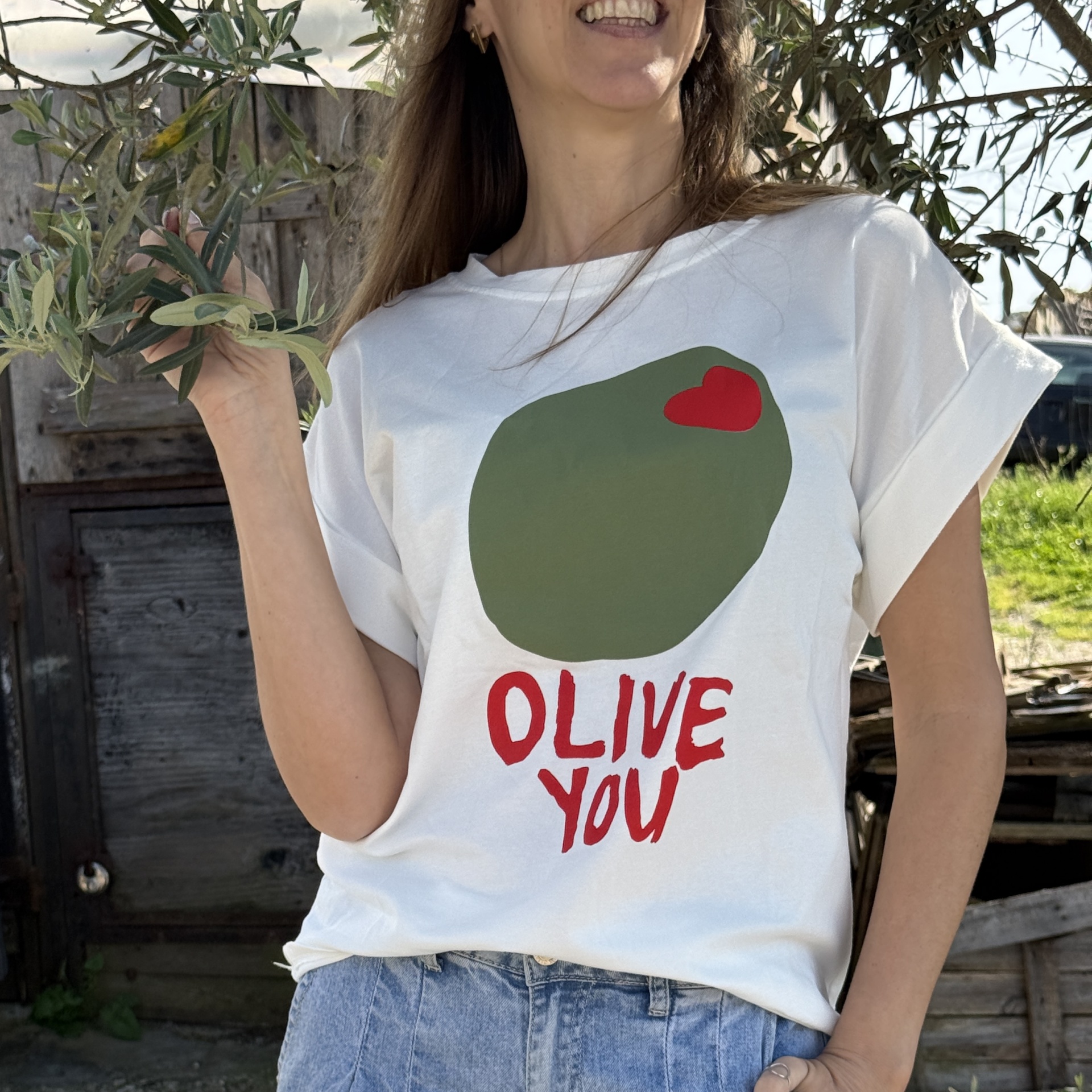 T-shirt branca com estampa verde e vermelha OLIVE YOU usada por mulher perto de uma oliveira