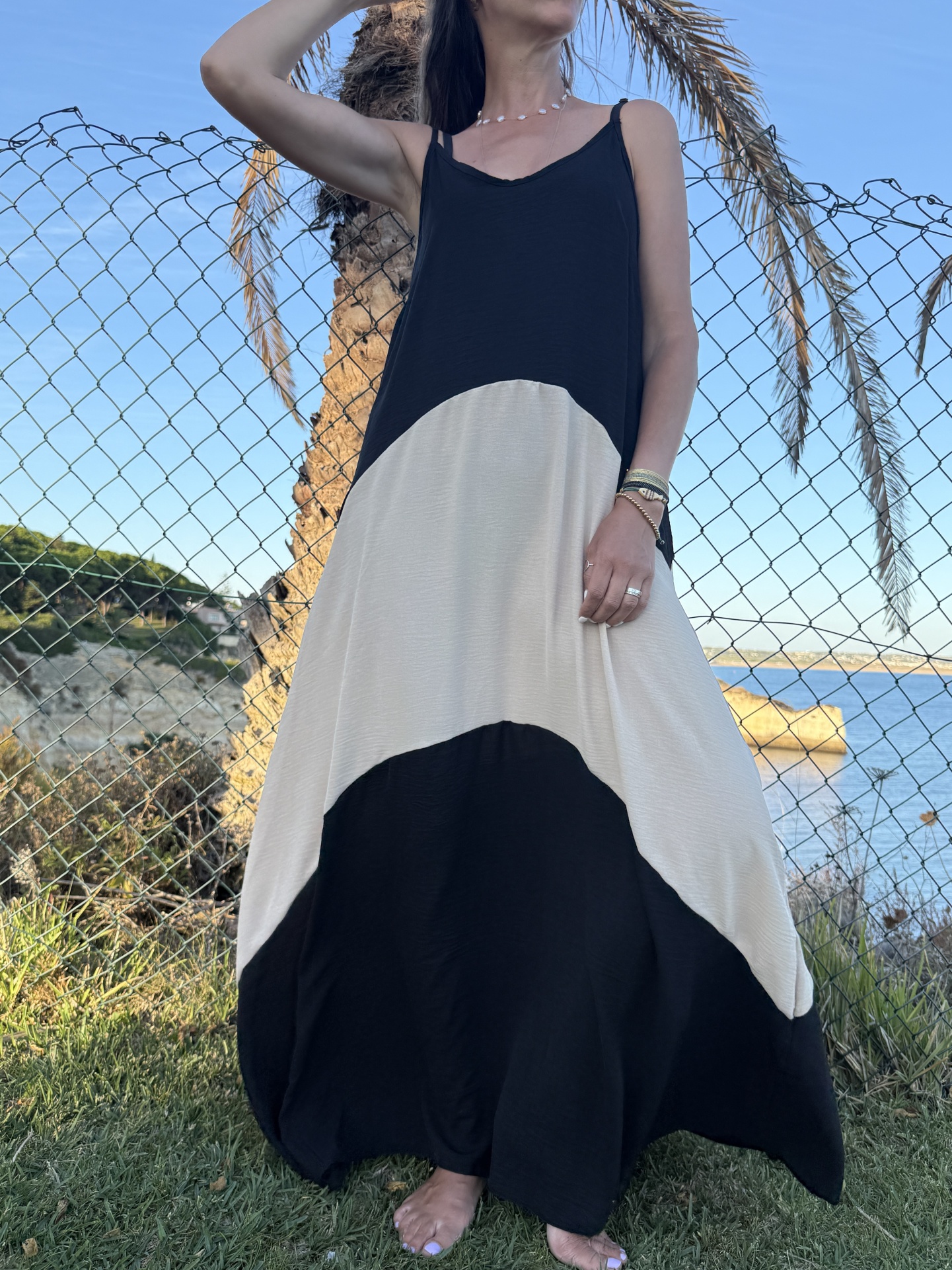 Vestido bicolor comprido em preto e bege claro, mulher ao ar livre perto de uma cerca de arame e mar
