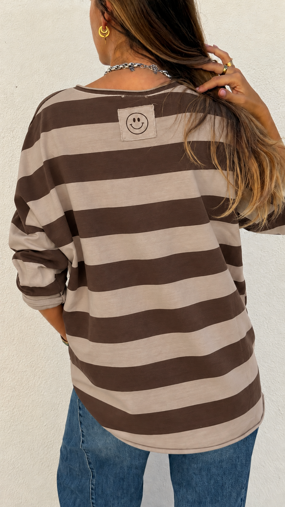 T-shirt riscas castanhas e bege com patch smiley nas costas, mulher vista de costas, acessórios dourados e colar prateado