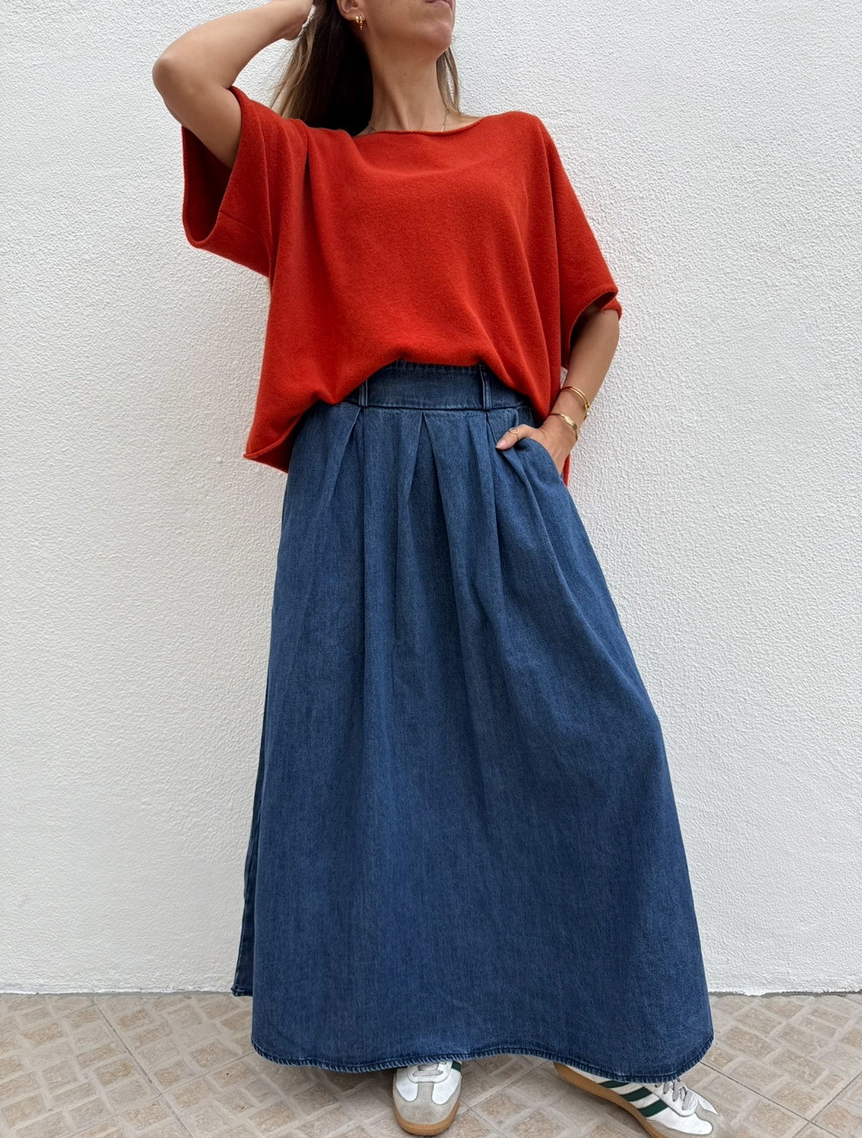 Mulher com camisola vermelha e saia comprida azul escura de denim junto a parede branca