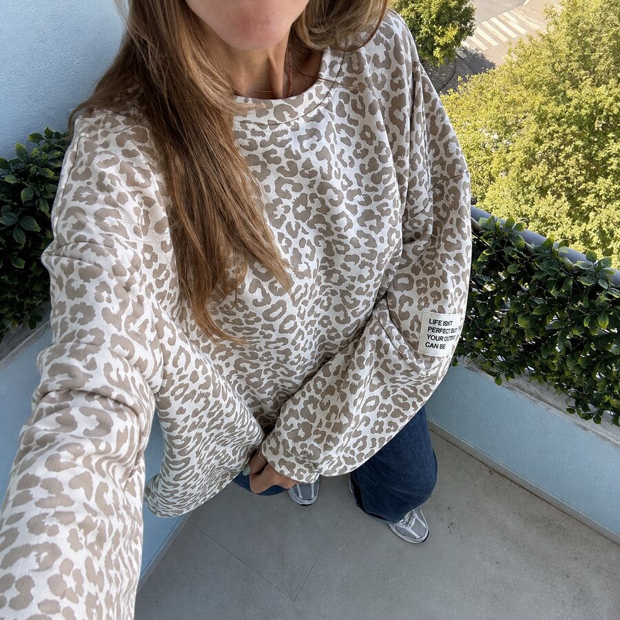 Sweatshirt bege com padrão animal print branco e etiqueta com texto na manga, usada numa varanda com plantas