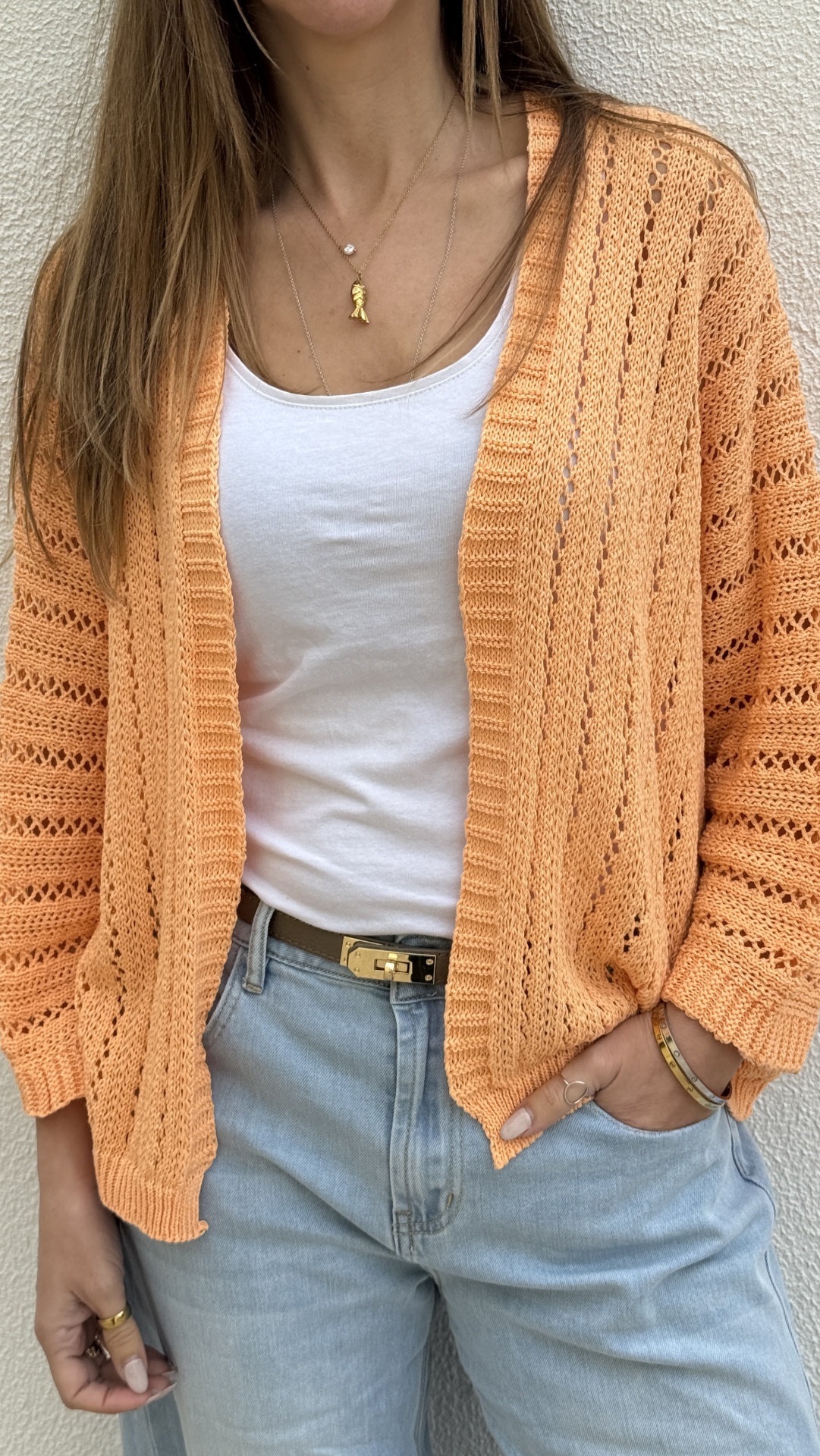 Mulher com casaco de malha pêssego, t-shirt branca, calças de ganga azuis e acessórios dourados