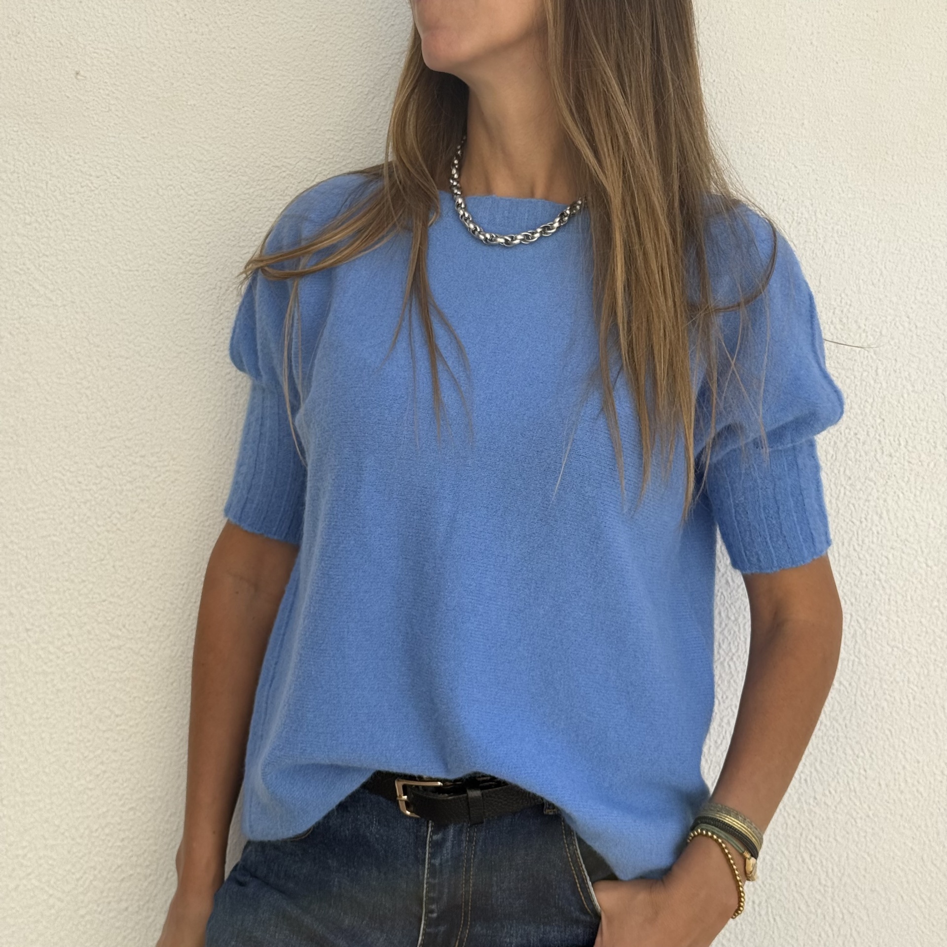 Camisola azul de malha, jeans escuros, colar prateado e pulseiras com fundo branco.