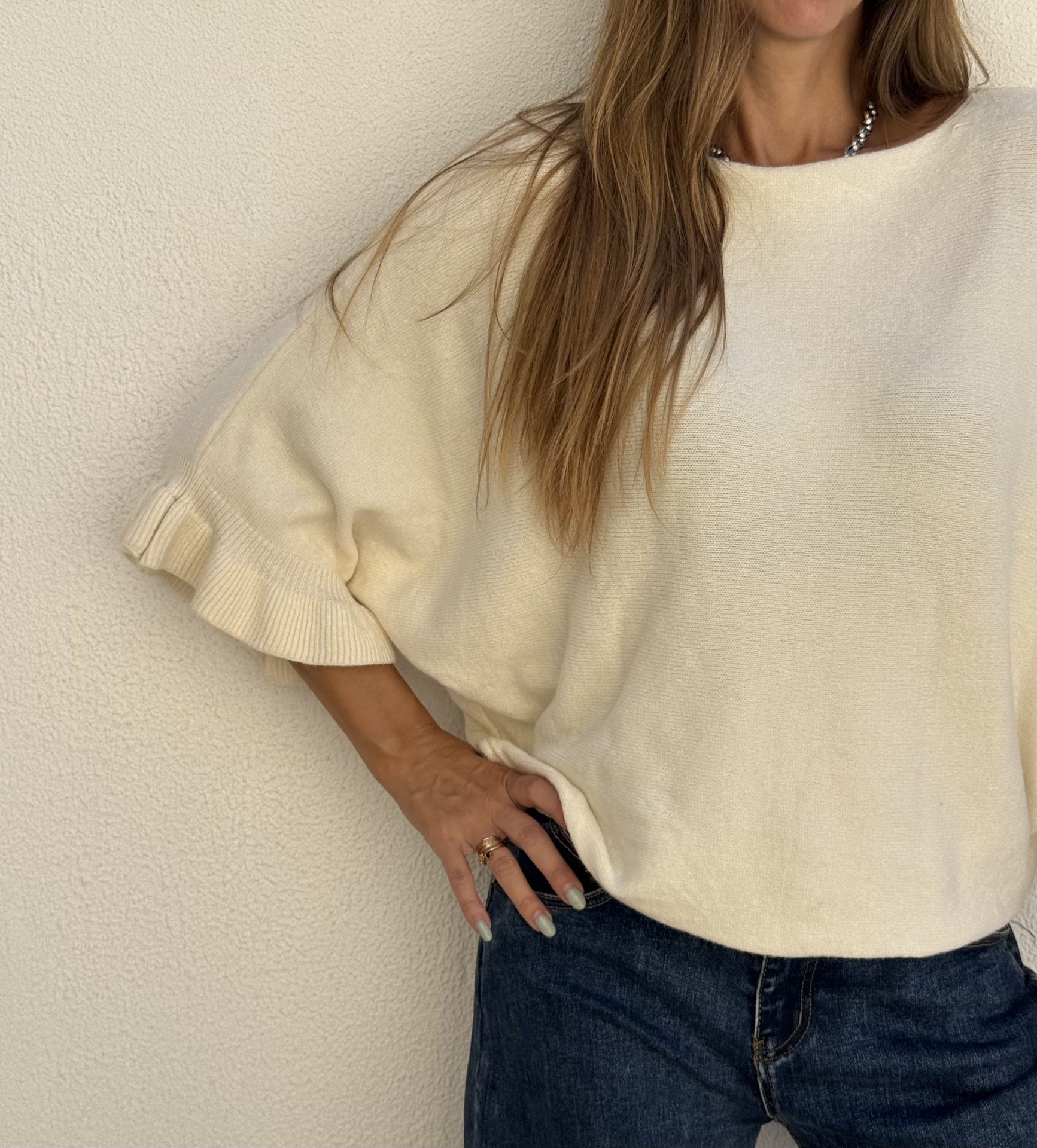 Camisola bege de malha com mangas de folho e calças de ganga escuras.