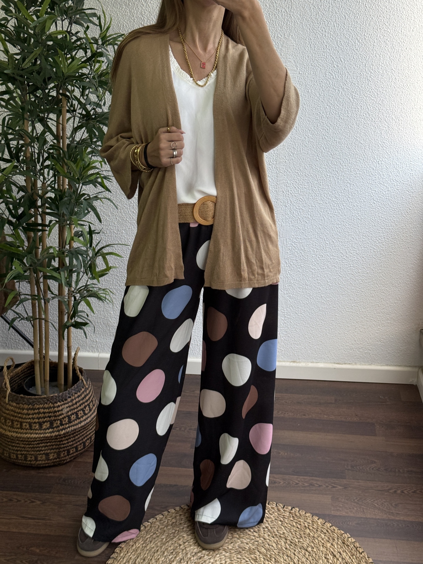 Mulher com cardigan bege, blusa branca, calças pretas com pintas coloridas e acessórios dourados, tirando selfie em espaço interior com planta ao lado.