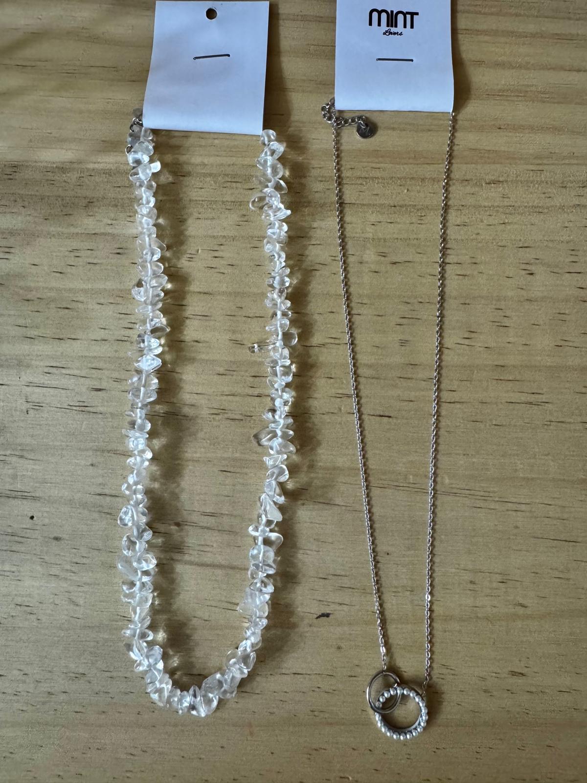Dois colares, um de pedras transparentes e outro com corrente prateada e pendente de argolas, sobre mesa de madeira
