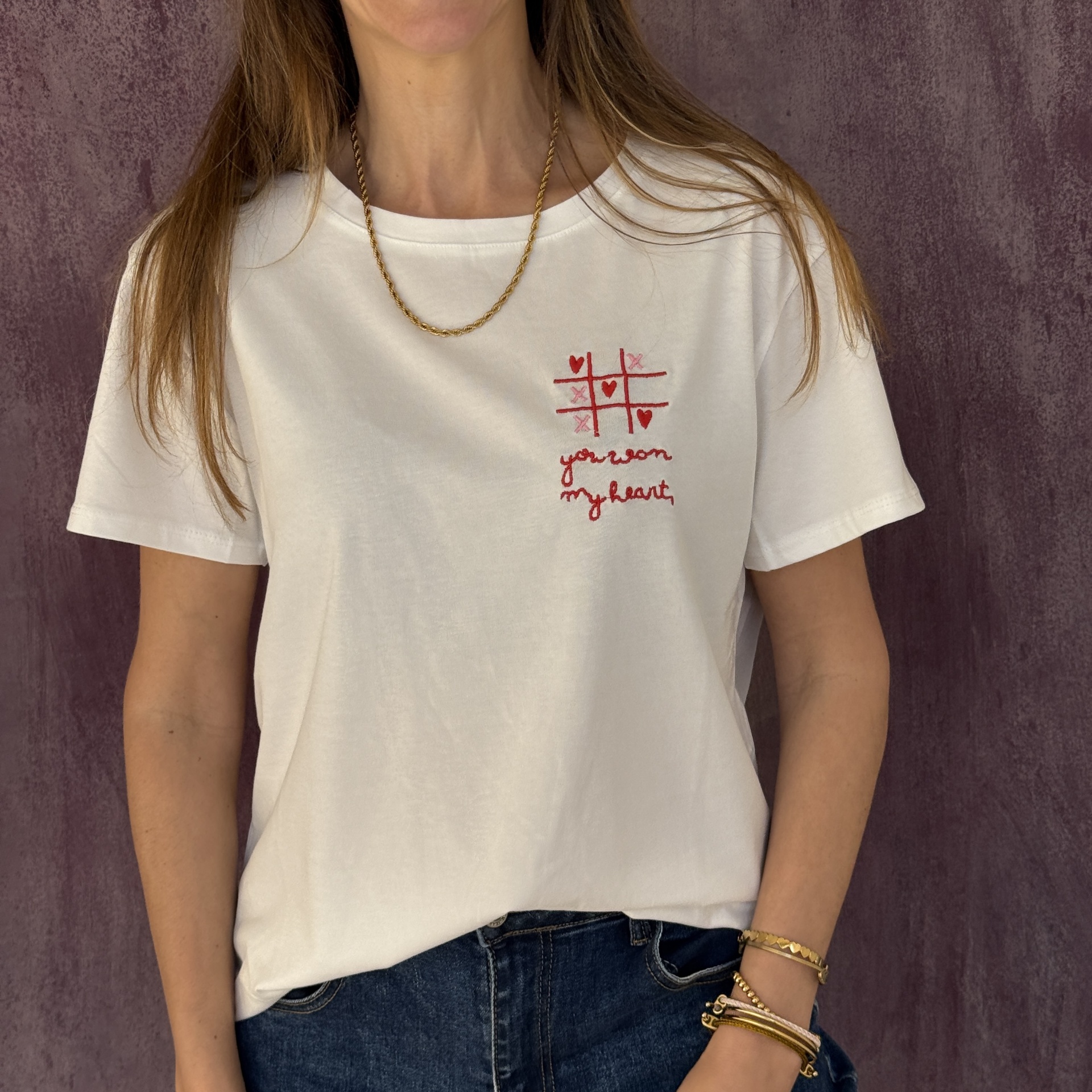 T-shirt branca com bordado de jogo-da-velha e texto you won my heart, usada com jeans azul escuro e pulseiras.