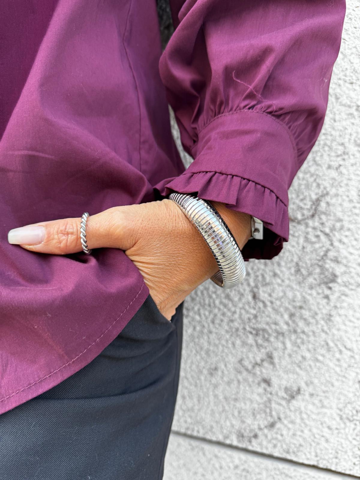 Mão com anel e pulseira prateados, camisa roxa com punho franzido, calças pretas, fundo cinzento.