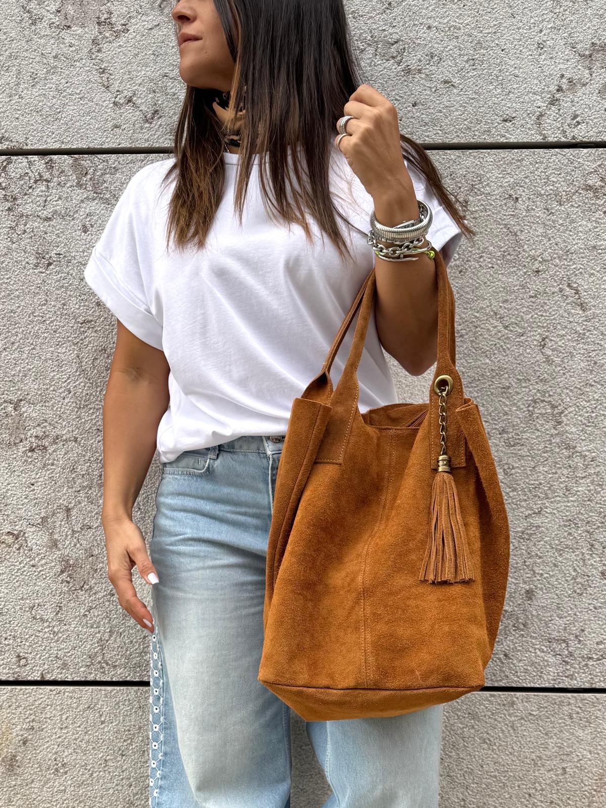 Mala castanha de camurça com tassel pendurado, usada por mulher com camisola branca e jeans azul claro, parede cinzenta ao fundo