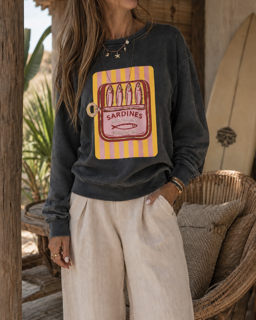 Sweatshirt preta com estampa colorida de lata de sardinhas, calças largas bege, cadeira de vime e prancha de surf ao fundo