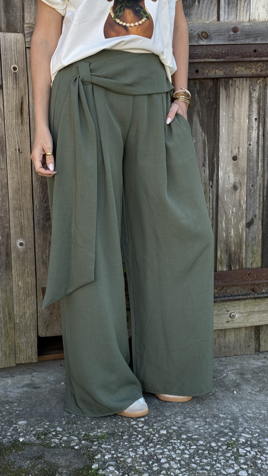 Calças largas verdes com cinto largo usada por pessoa com pulseiras e anel junto a porta de madeira