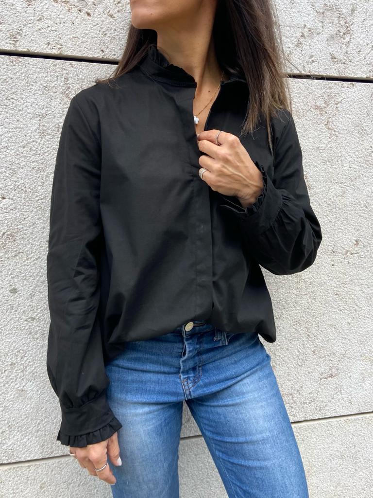 Blusa preta com gola alta e punhos com folhos, usada com jeans azuis, em fundo de parede cinzenta.