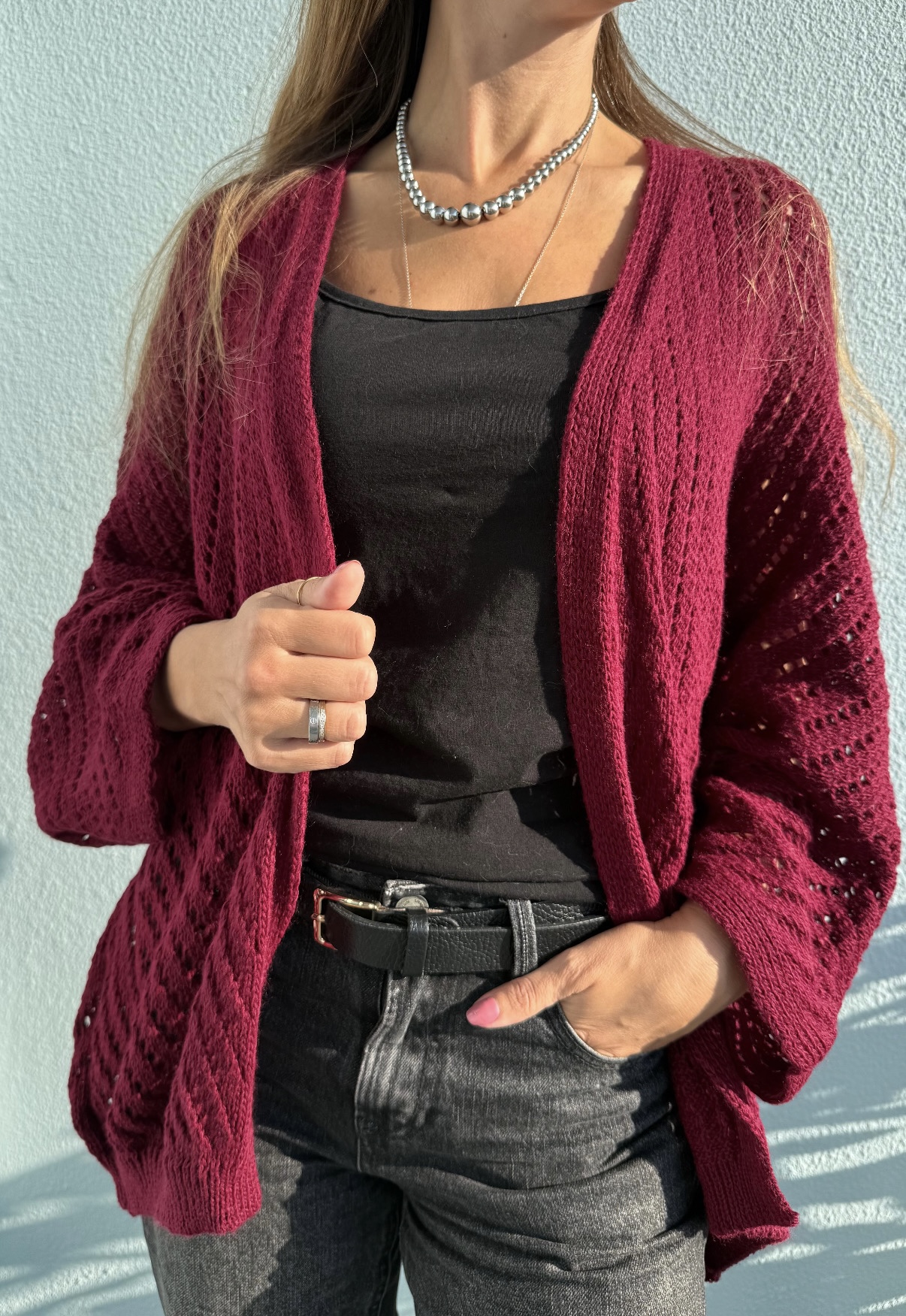 Casaco vinho de malha, camisola preta, calças jeans cinzentas e acessórios prateados em modelo