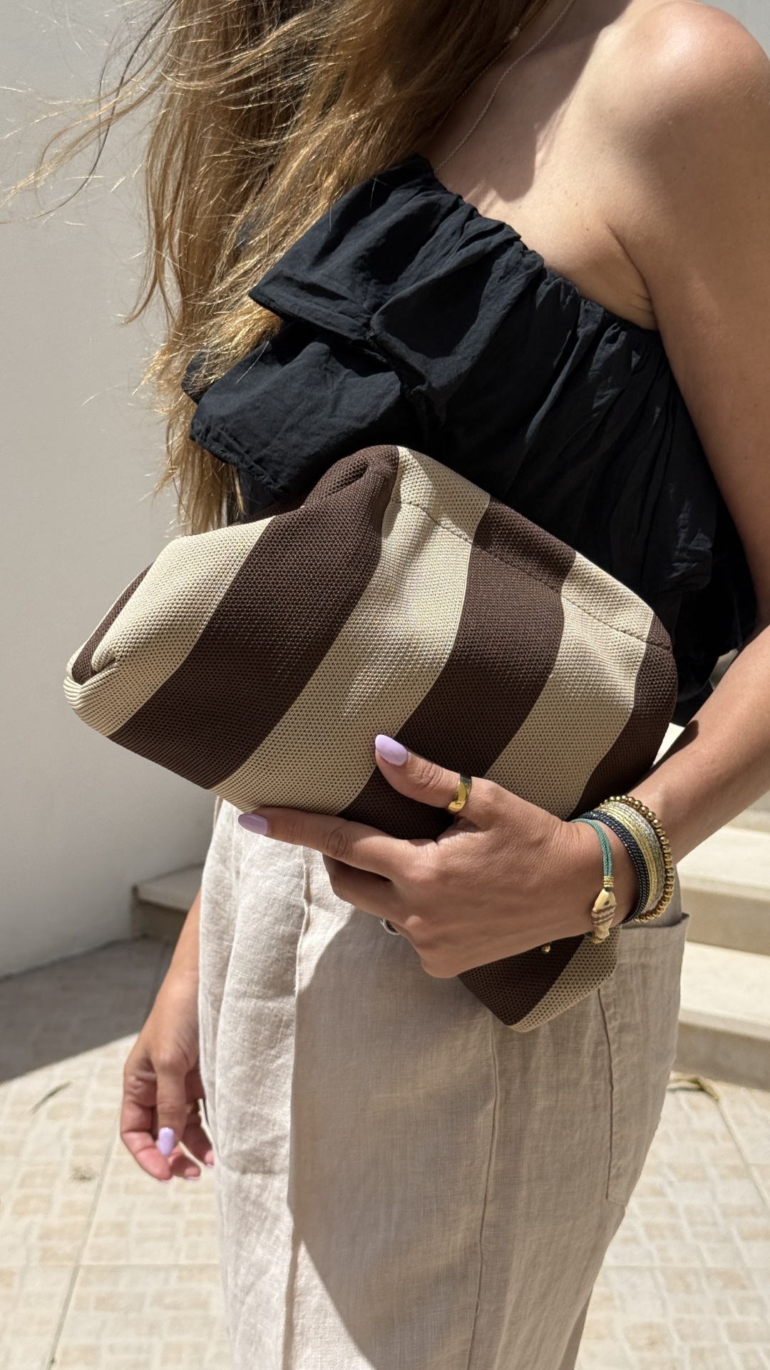 Mala clutch riscada castanha e bege em mão de mulher com blusa preta e calças bege