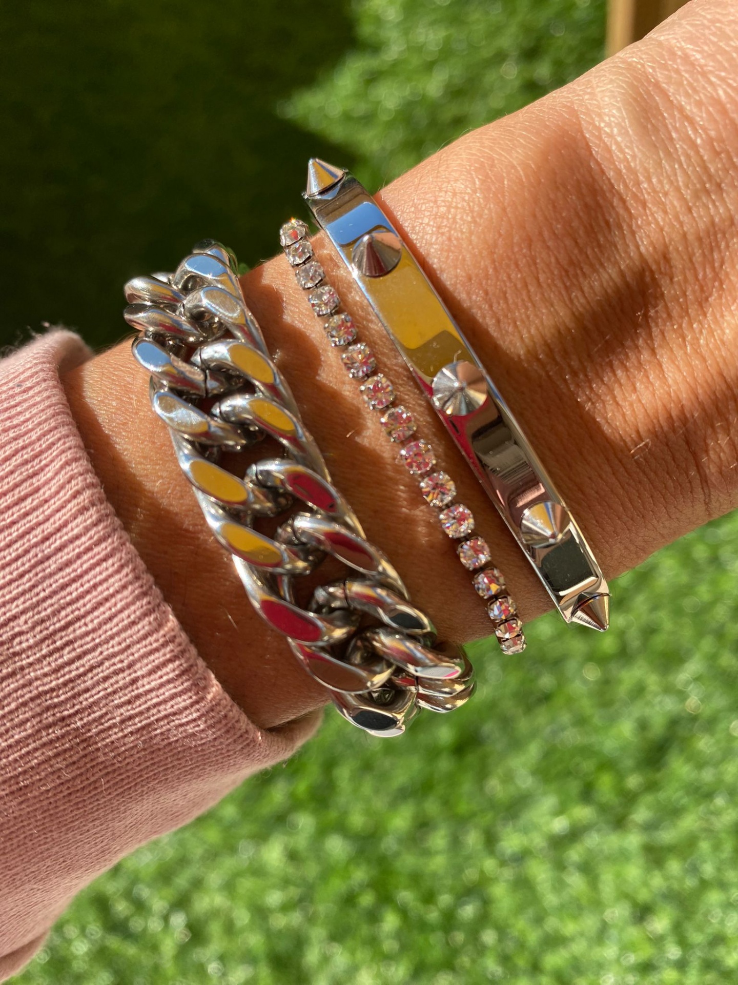 Três pulseiras prateadas no pulso, uma com elos grossos, outra com pedras, e outra com tachas e parafusos, com fundo de relva.