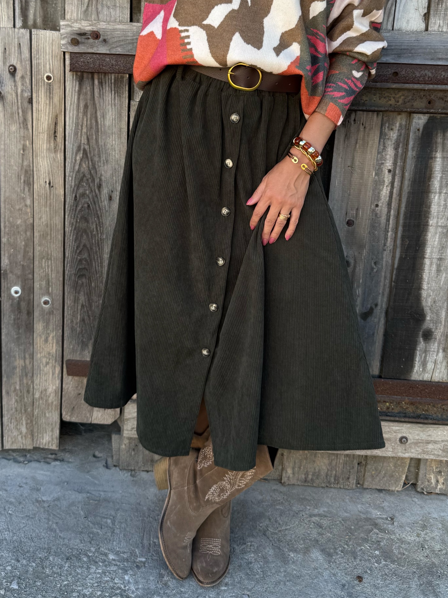 Saia midi preta com botões, cinto castanho, botas castanhas bordadas, suéter colorido, pulseiras, fundo madeira