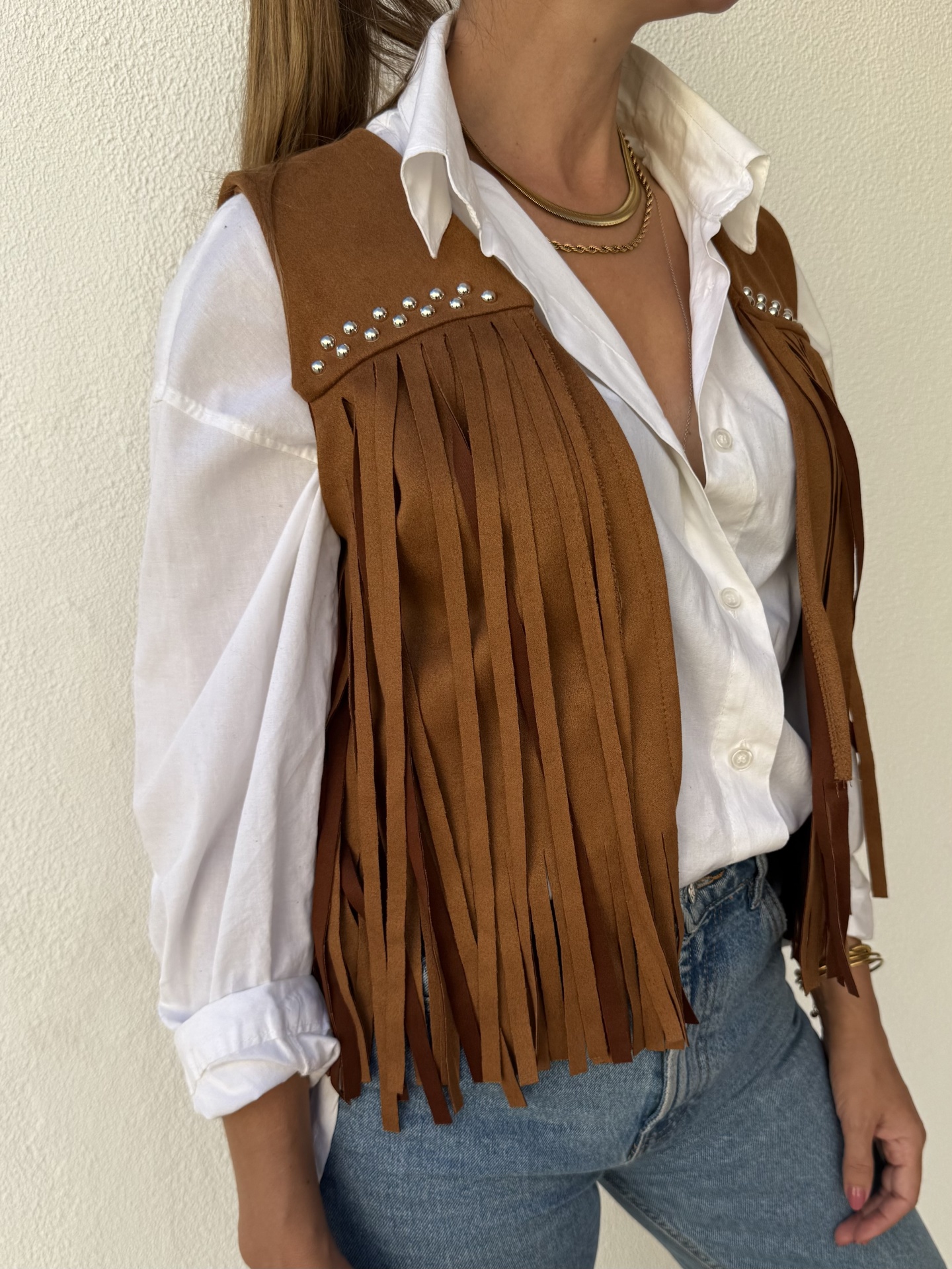 Colete castanho com franjas e tachas sobre camisa branca e jeans azuis