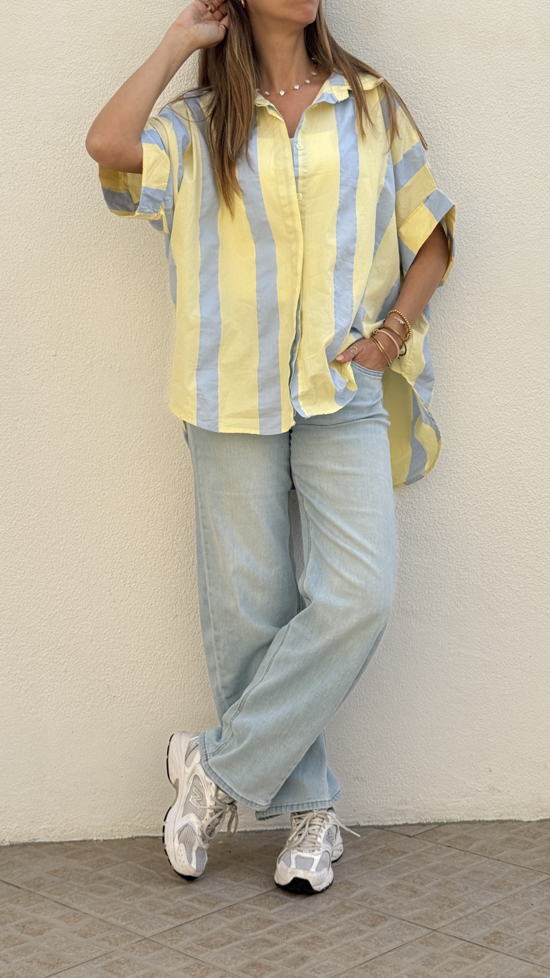 Pessoa vestindo blusa de riscas amarelas e azuis, calças de ganga claras e ténis brancos, em chão de azulejos.