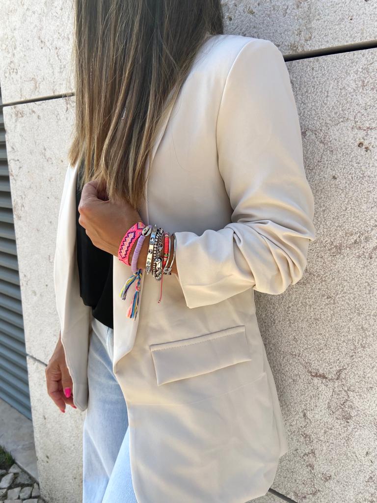 Blazer bege claro com pulseiras coloridas no pulso e calças azuis claras junto à parede