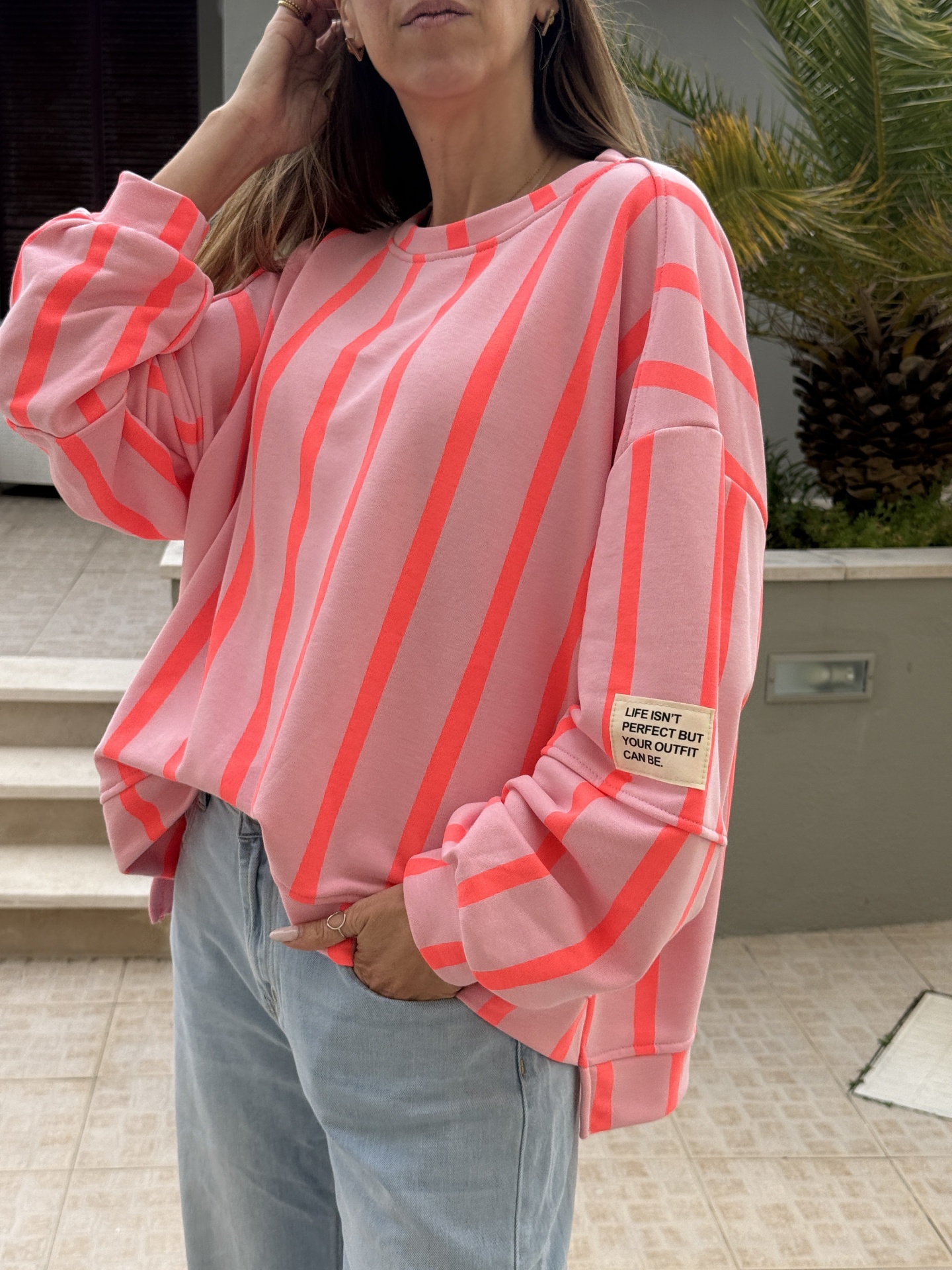 Sweatshirt rosa com riscas verticais cor de salmão e etiqueta na manga, usada com jeans azuis claros.