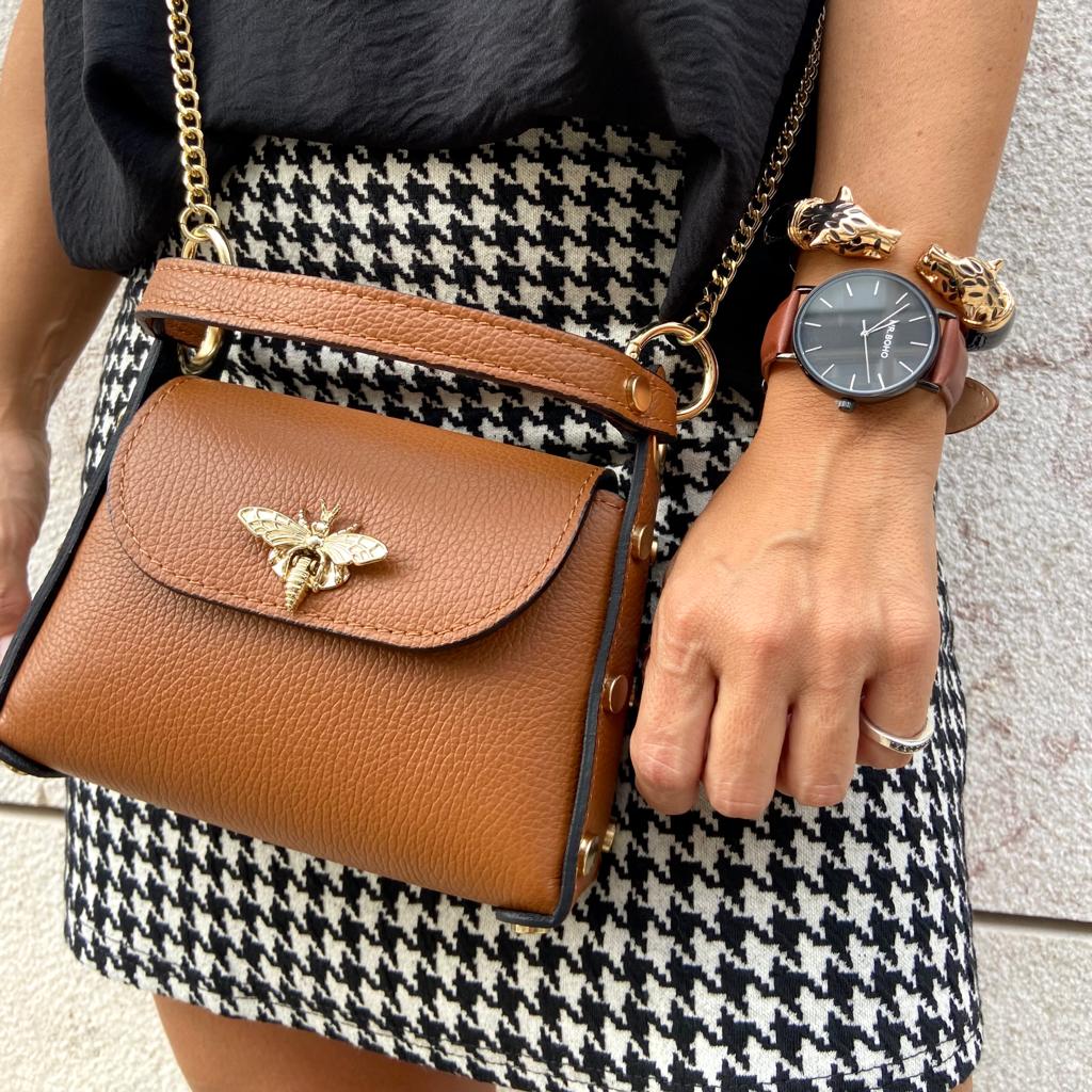 Bolsa castanha em couro com corrente dourada, pulseira e relógio no braço de mulher com saia pied-de-poule preta e branca