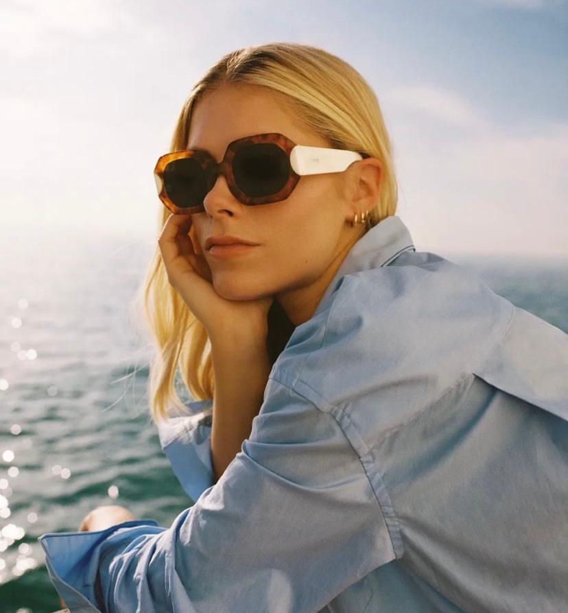 Mulher loira com óculos de sol castanhos e brancos junto ao mar vestindo camisa azul clara