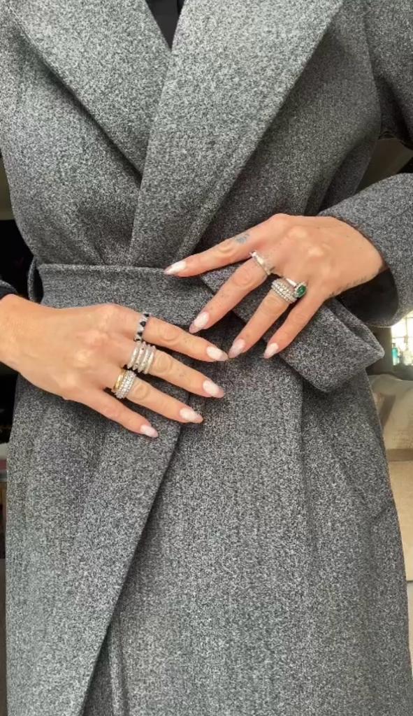 Mulher a usar casaco cinza com cinto, mãos com anéis variados.