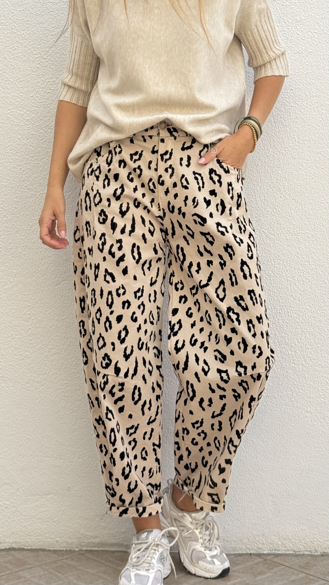 Pessoa com calças animal print bege e camisola bege posando numa parede branca.
