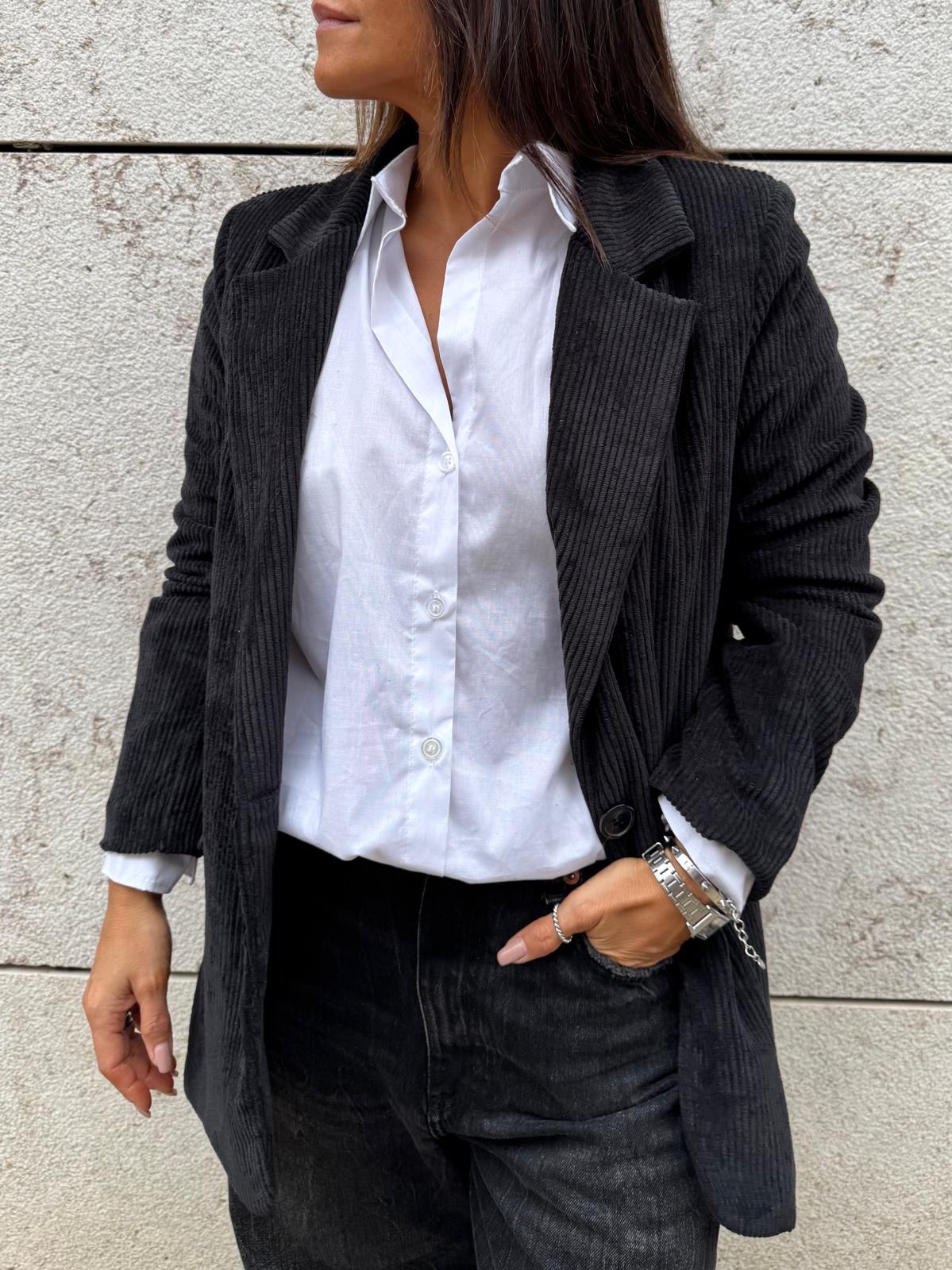 Mulher com camisa branca, blazer preto de tecido canelado e calças pretas, pulseiras e anel, diante de parede clara texturizada