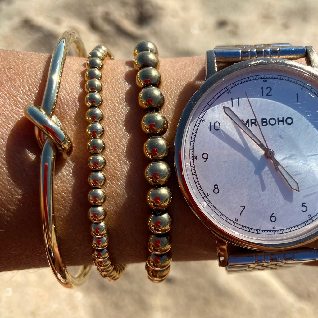 Pulso com relógio MR.BOHO e três pulseiras douradas sobre fundo desfocado. 